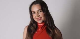 Smiling woman with long brown hair wearing a glossy red sleeveless top, posing against a light gray backdrop.