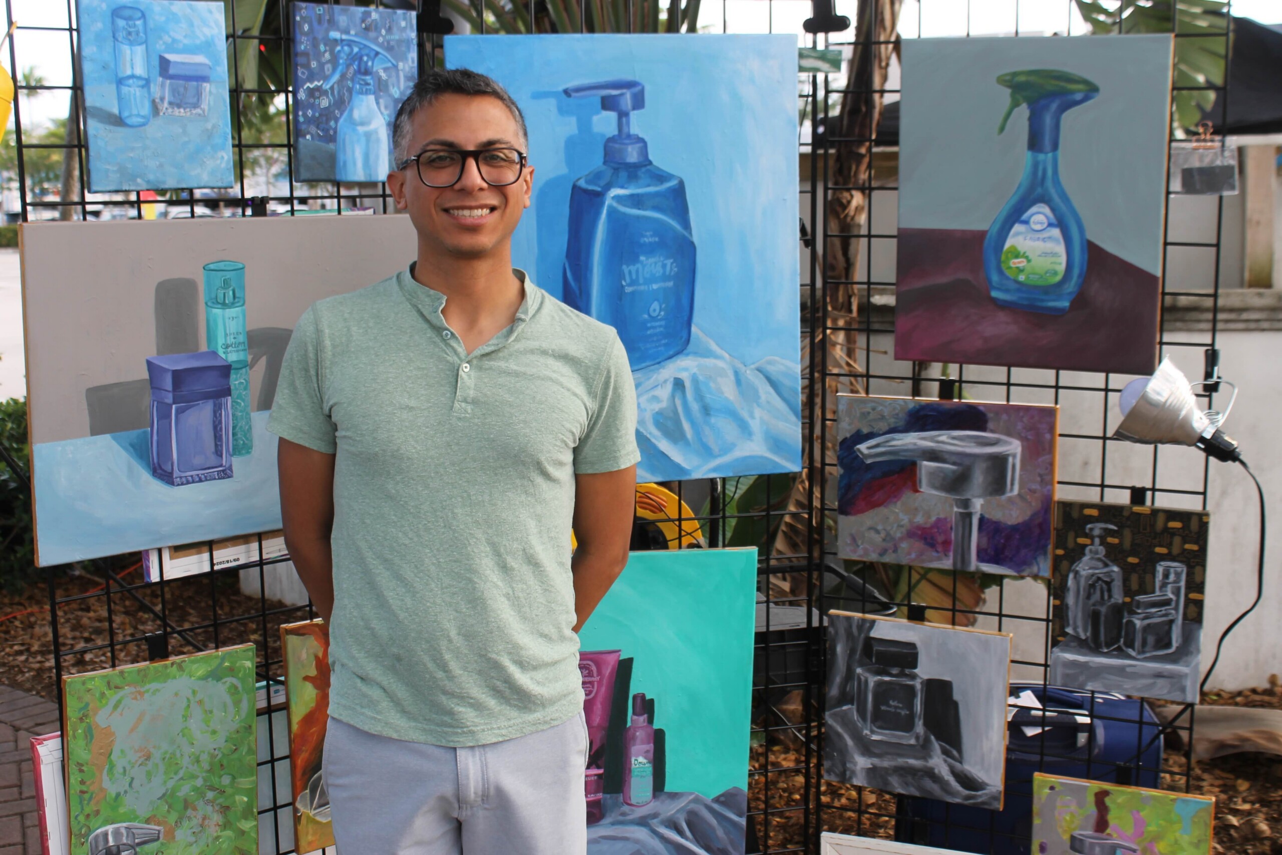 Smiling man in a mint T-shirt stands in front of an outdoor art display of blue bottle paintings on a grid fence.