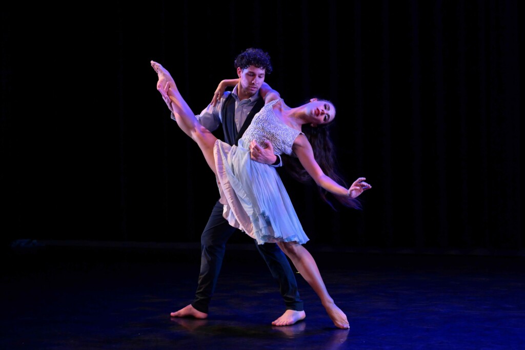 Dance couple performing a dramatic lift on a dark stage; the woman extends a leg while the man supports her.