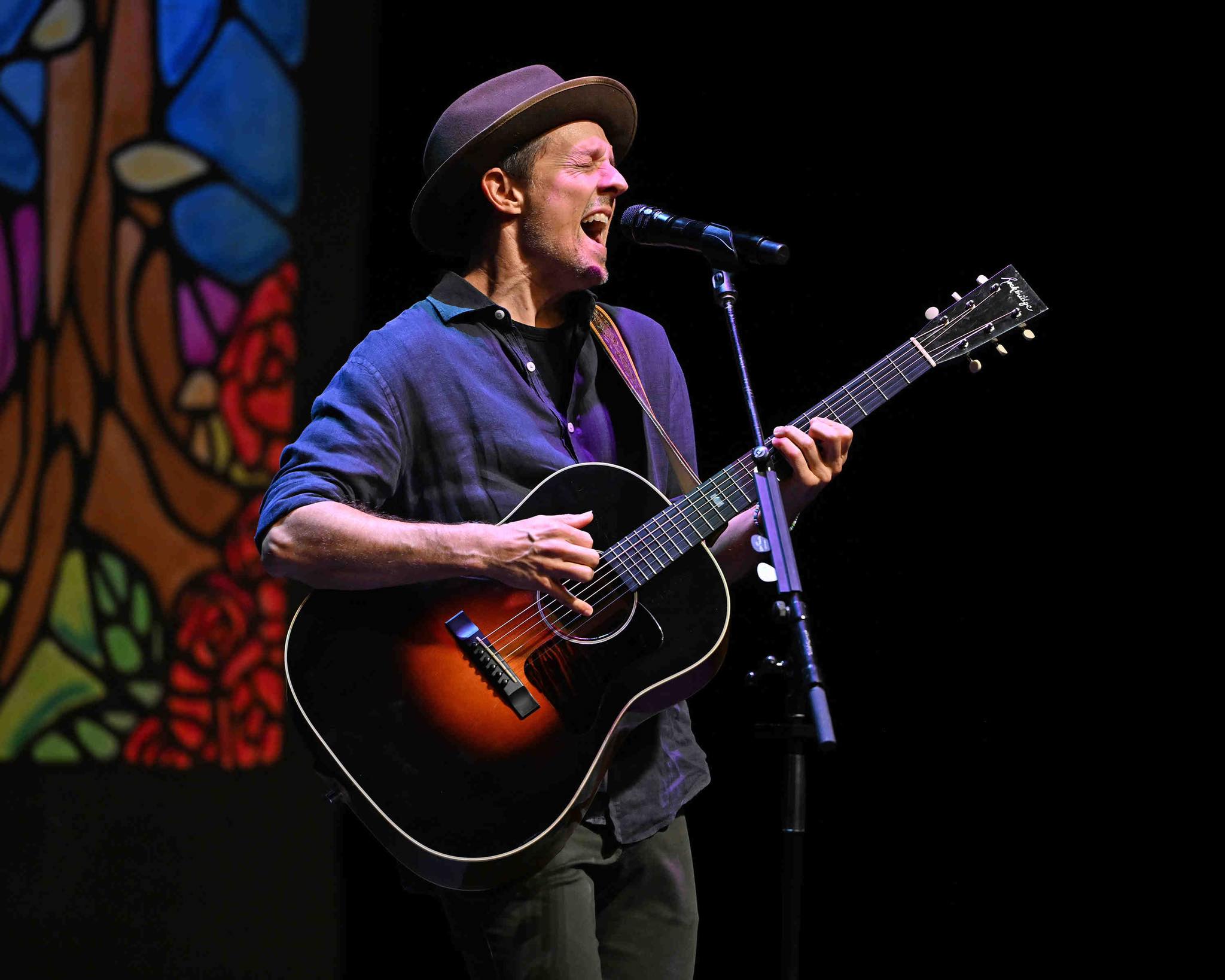 Male musician singing into microphone while playing an acoustic guitar on stage; wearing a hat, blue shirt, and carrying a strap against a dark background.