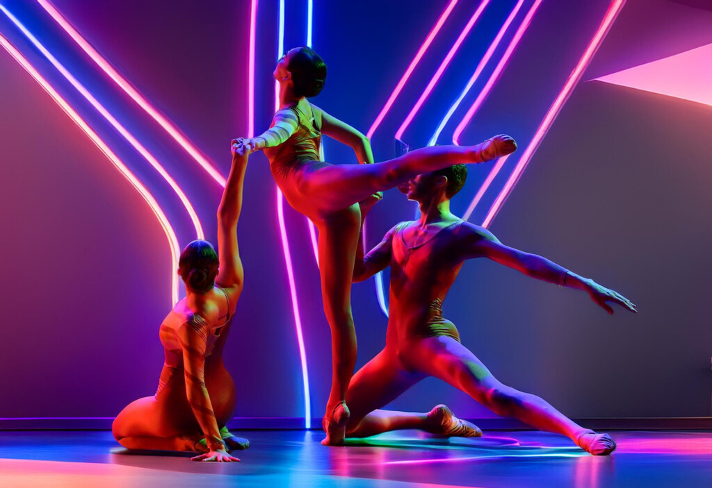 Dancers perform a high lift on a neon-lit stage, pink and blue tubes glowing around them.