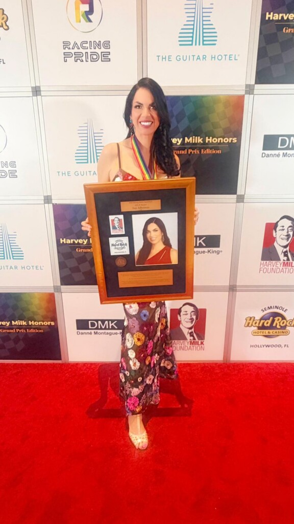 Woman on a red carpet holding a framed award, smiling at camera, wearing a floral dress and medal necklace.
