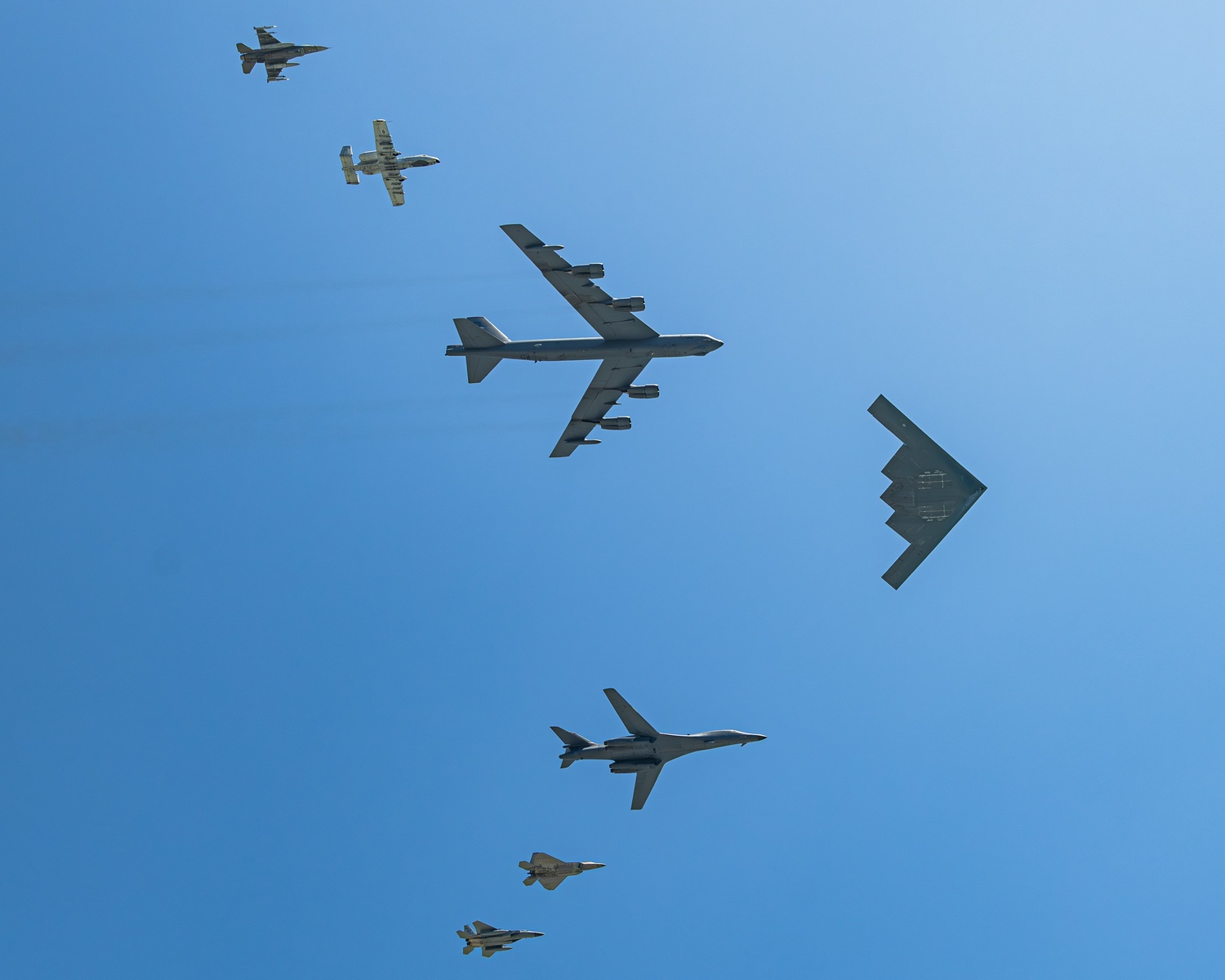 Formation of military aircraft in clear blue sky: a large B-52 bomber, a B-2 Spirit stealth bomber, and several fighters fly in formation.