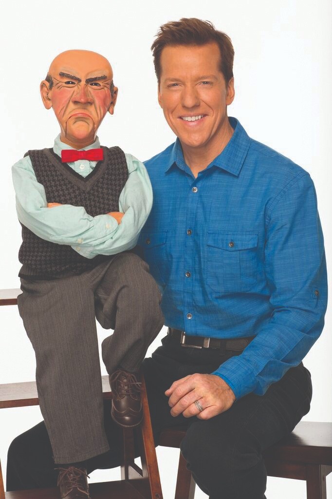Ventriloquist smiling beside a stern-looking puppet wearing a vest and red bow tie, seated on a bench.