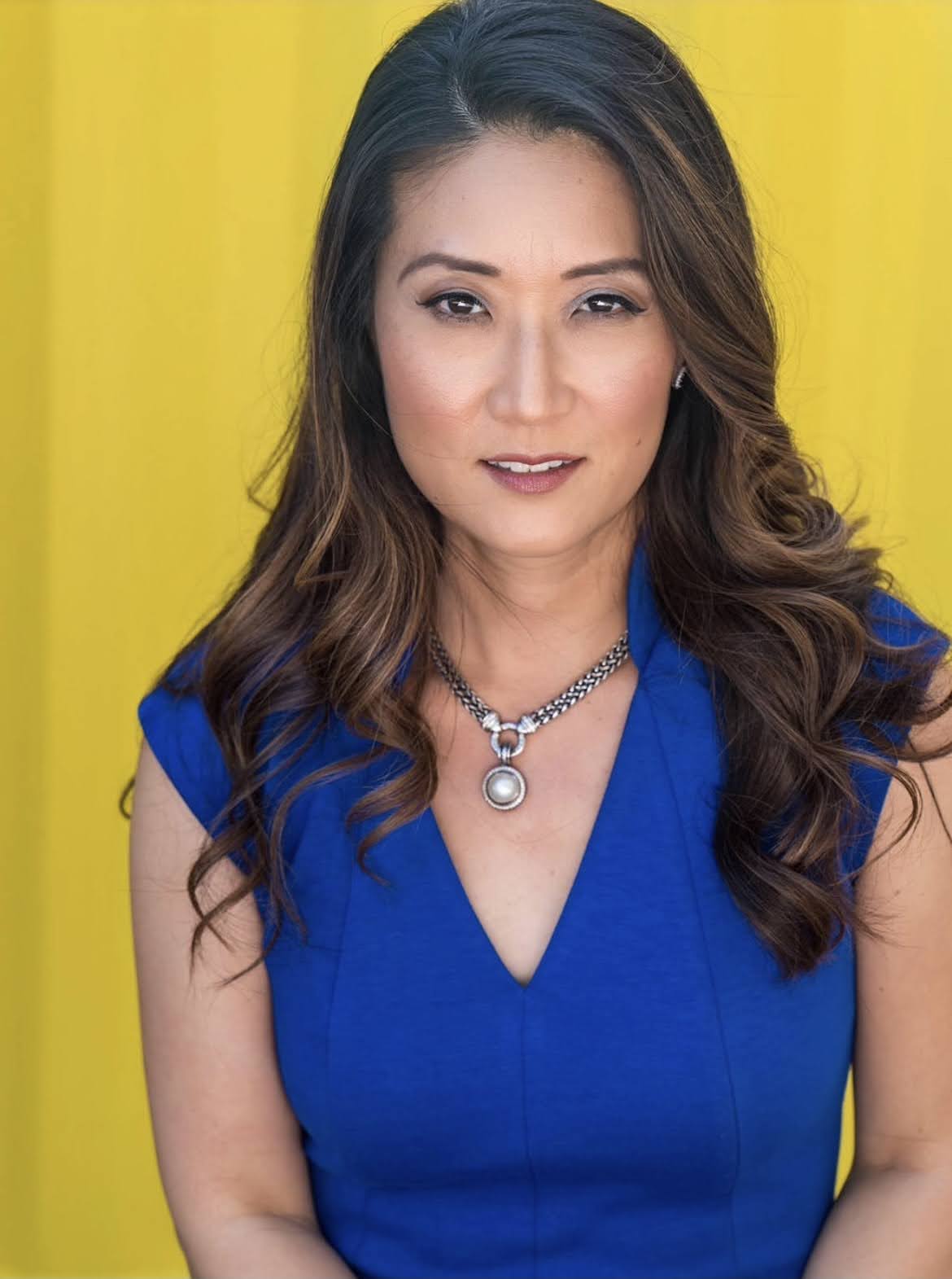 2nd Annual Pride Center Drag Brunch: At Seminole Hard Rock Hotel & Casino Hollywood | Hotspots Magazine Hotspots Magazine Portrait of a woman with wavy brown hair in a cobalt blue dress, wearing a silver pendant necklace, against a bright yellow background.