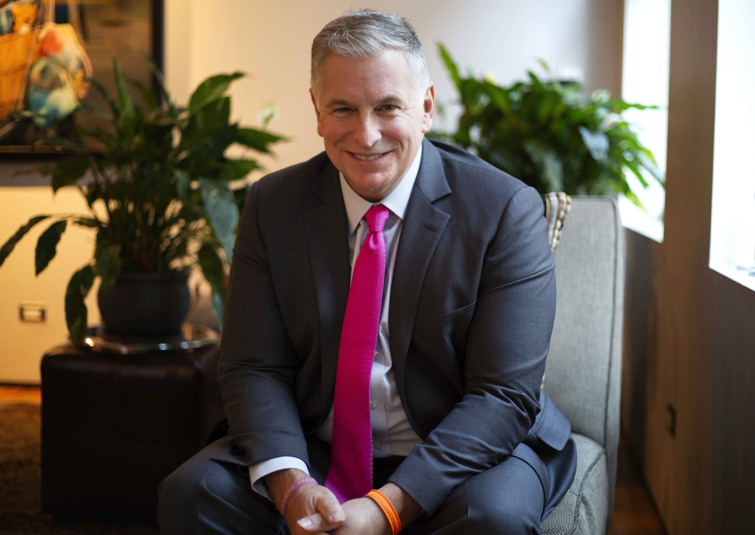 Smiling man in a dark suit and pink tie seated in an office with green plants in the background.