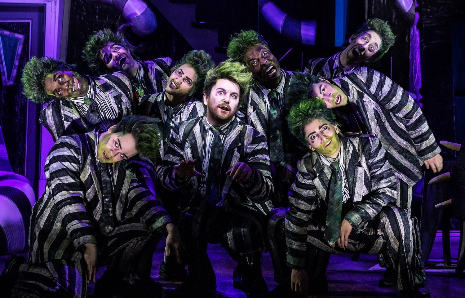 Group of performers in black-and-white striped suits with green makeup, posing together on a dimly lit stage.