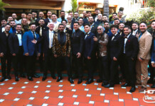 Large group of men in suits and a few women in formalwear pose on an orange-tiled terrace at a formal event.
