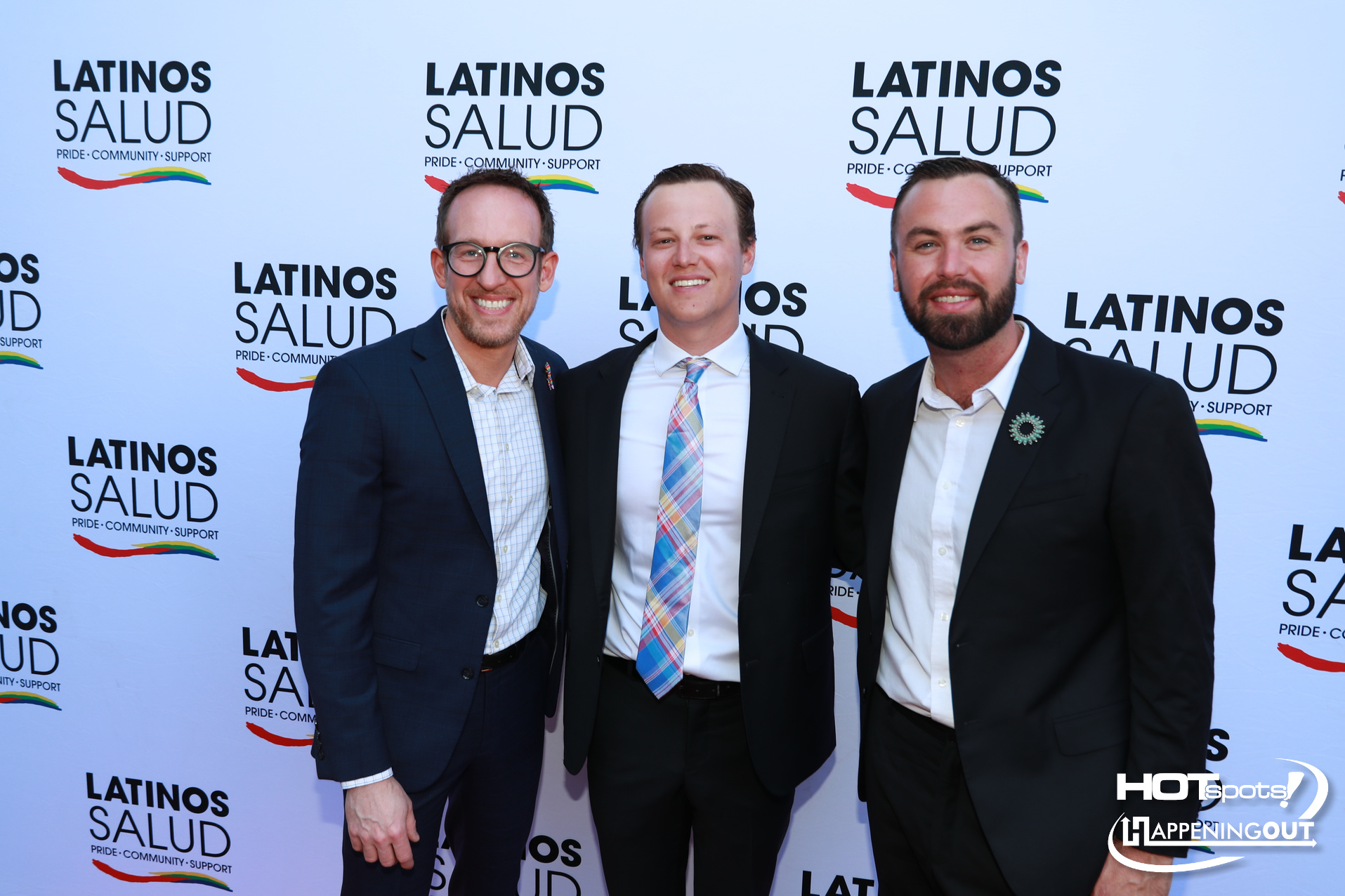 Three men in suits posing at a Latino Salud event with a step-and-repeat backdrop reading LATINOS SALUD and Pride • Community • Support