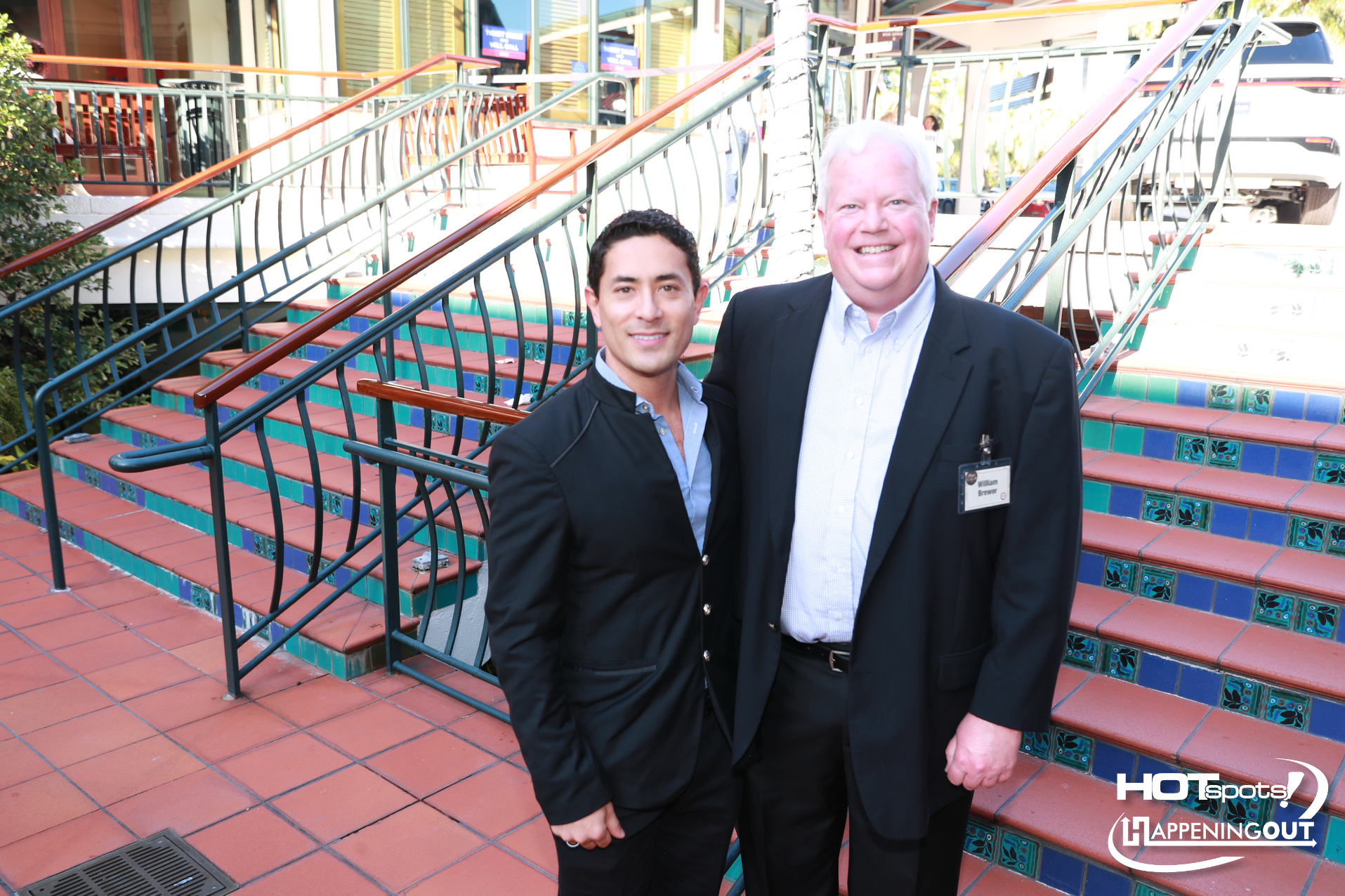 Two men in suits posing on colorful tiled stairs outdoors, one wearing a name badge; bright day.