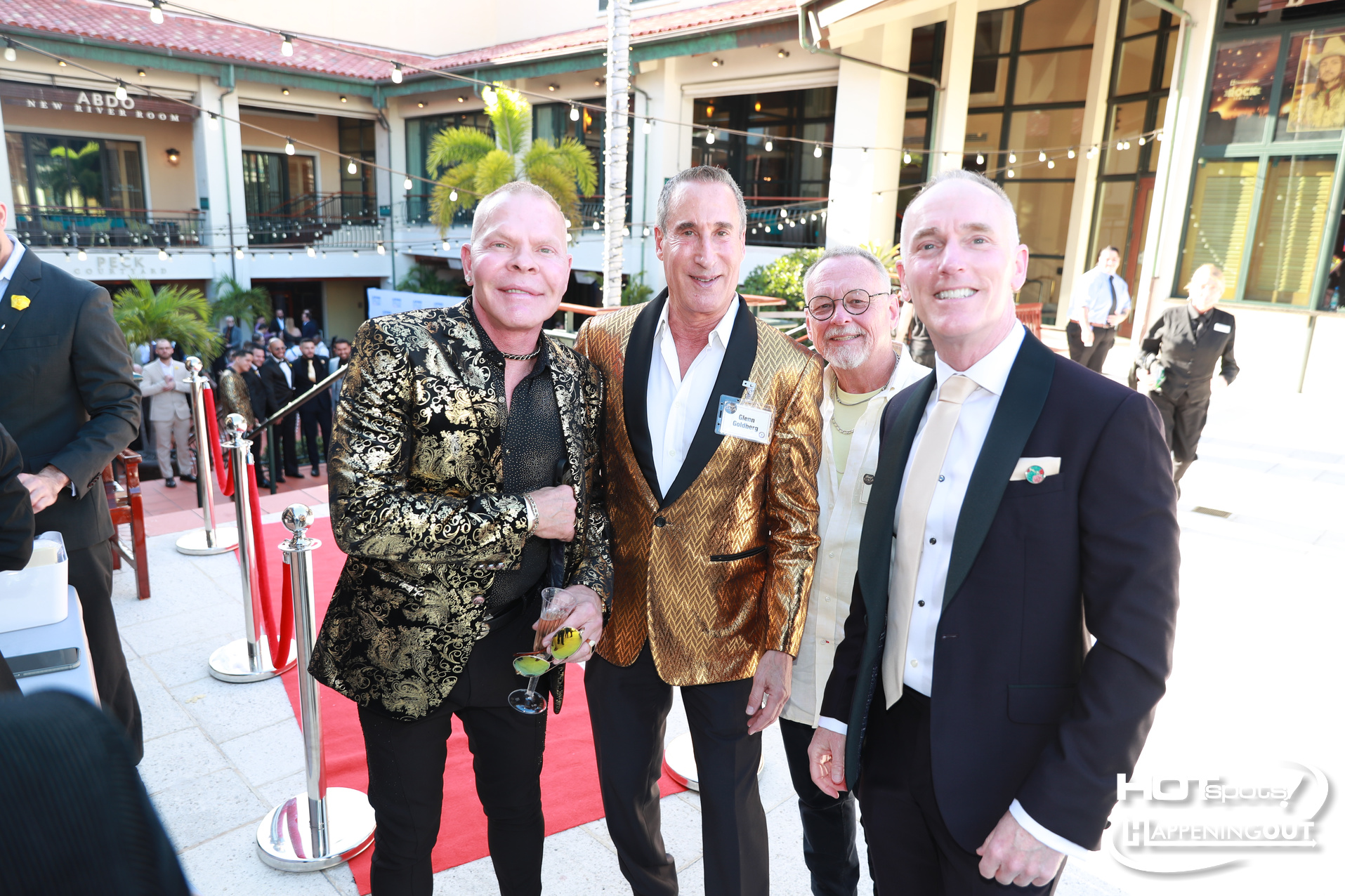 Four men in formal jackets stand on a red carpet at an outdoor event, string lights overhead and a crowd in the background.
