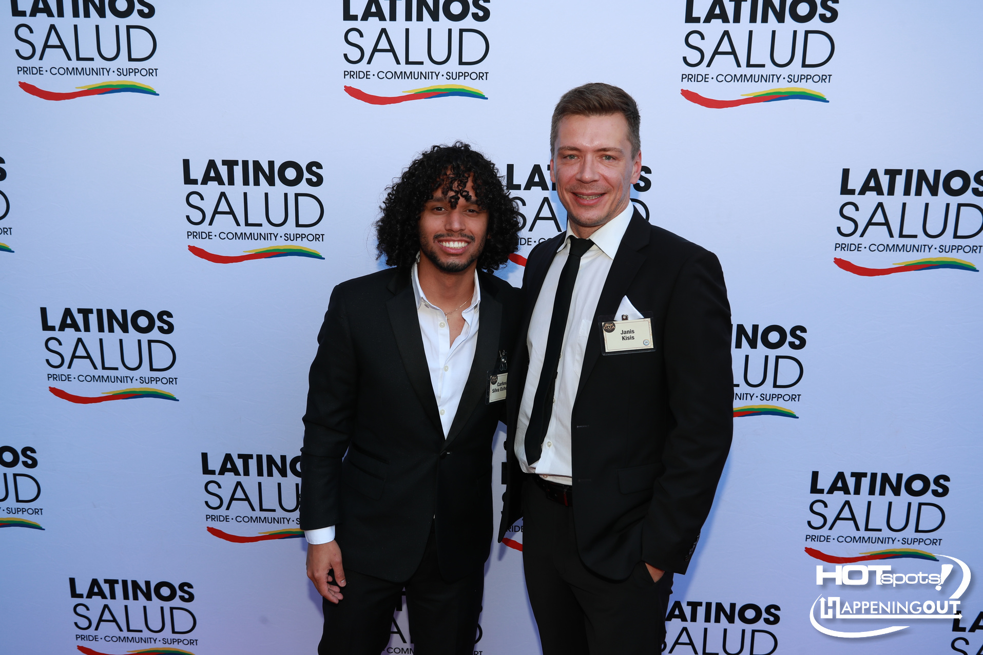 Two men in suits pose together at a Latinos Salud event backdrop with a rainbow logo and the words Pride, Community, Support in view.