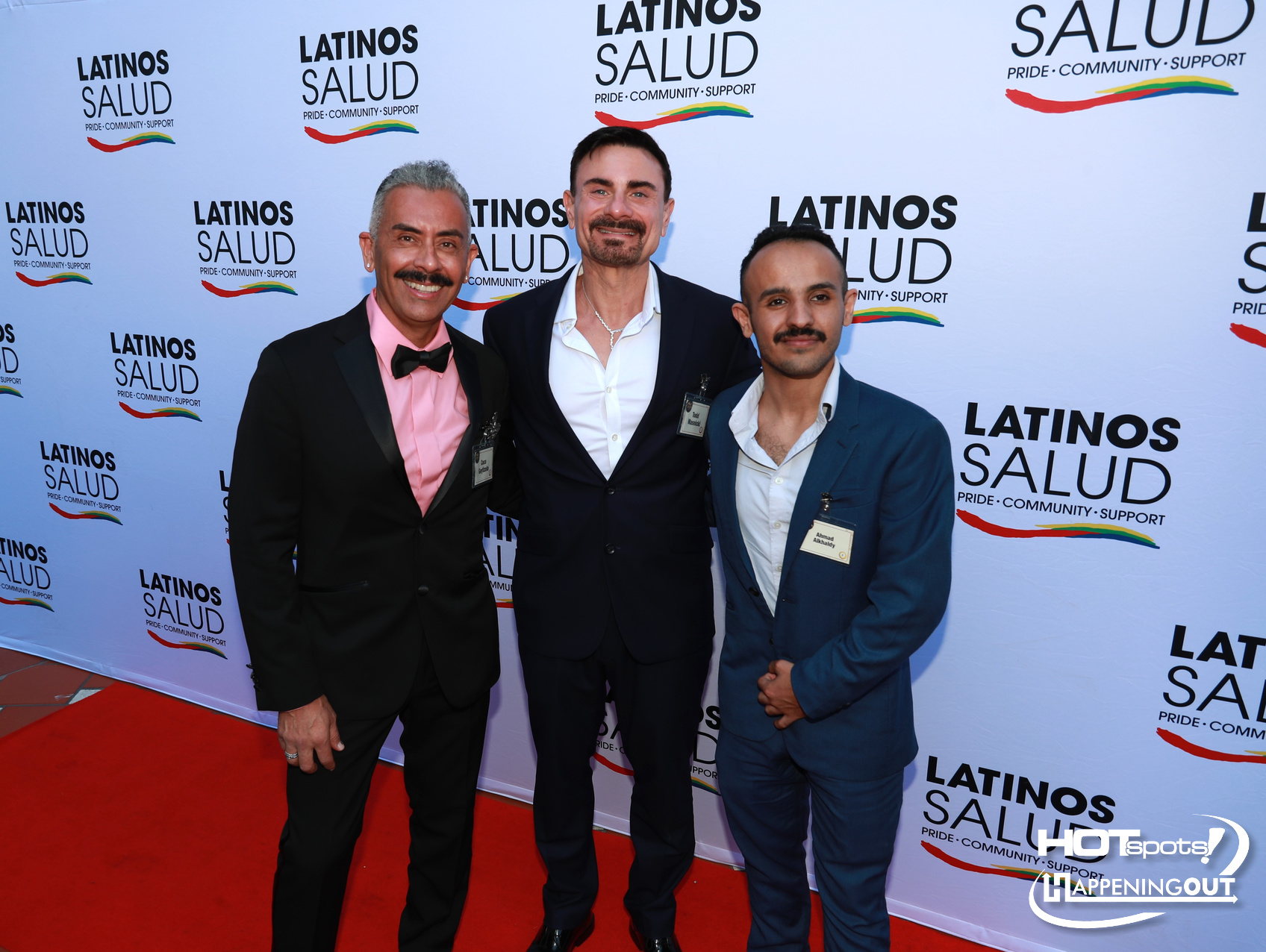 Three men in suits posing on a red carpet at a Latinos Salud event, with name badges and a backdrop covered in Latinos Salud logos.