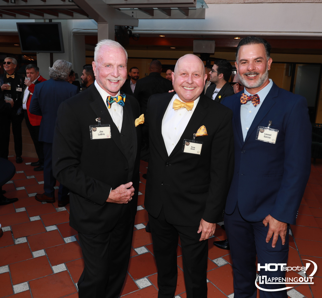 Three men in formal suits pose together at a social event, smiling and wearing bow ties and name badges.