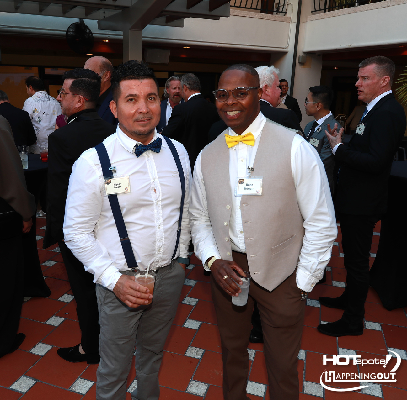 Two men in semi-formal attire pose for a photo at a cocktail event, each holding a drink and wearing name badges.