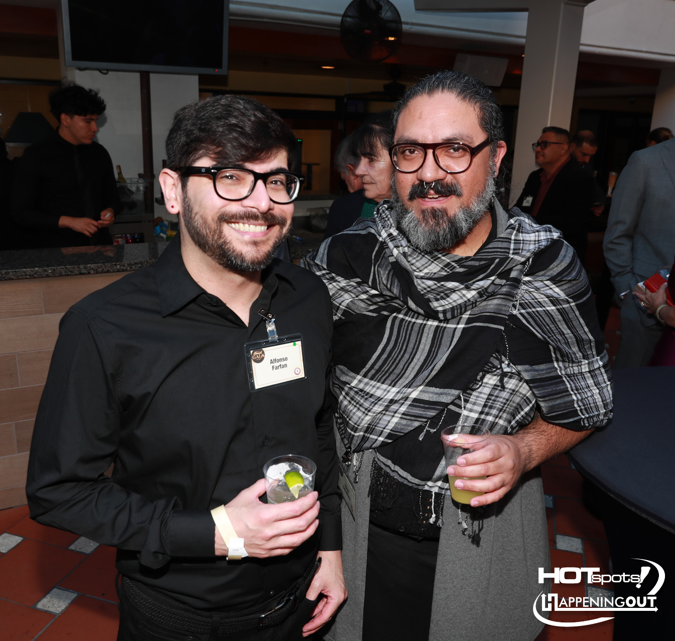 Two men smile for a photo at a party, each holding a drink; one in a black shirt with glasses, the other with a gray scarf and beard in front of a bar.