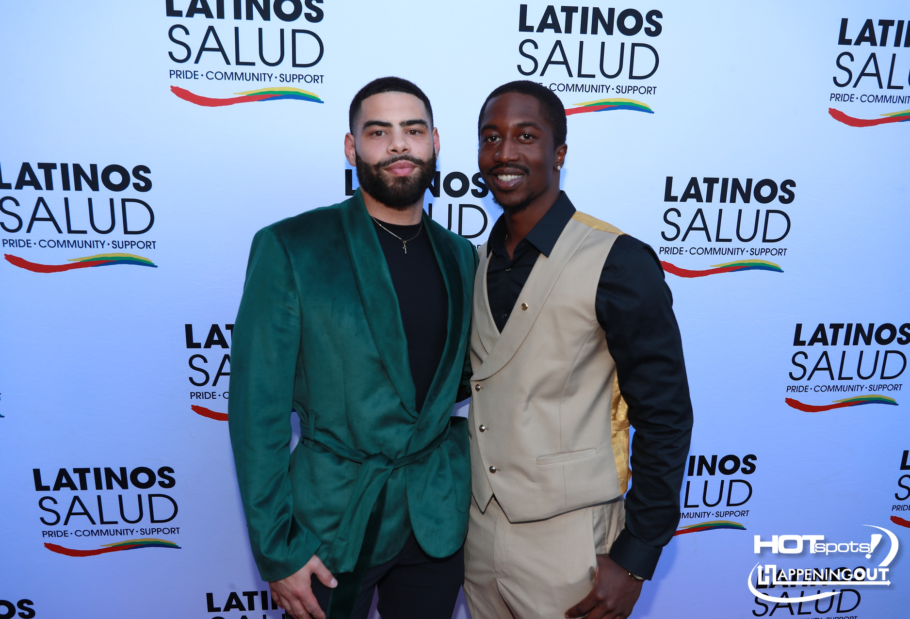 Two men pose together at a Latinos Salud event on a bannered backdrop; one wears a green velvet blazer, the other a beige vest and black shirt.