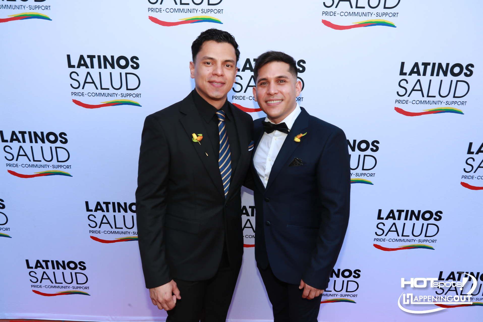 Two men in suits posing arm-in-arm at a Latinos Salud event backdrop with Pride colors logo behind them
