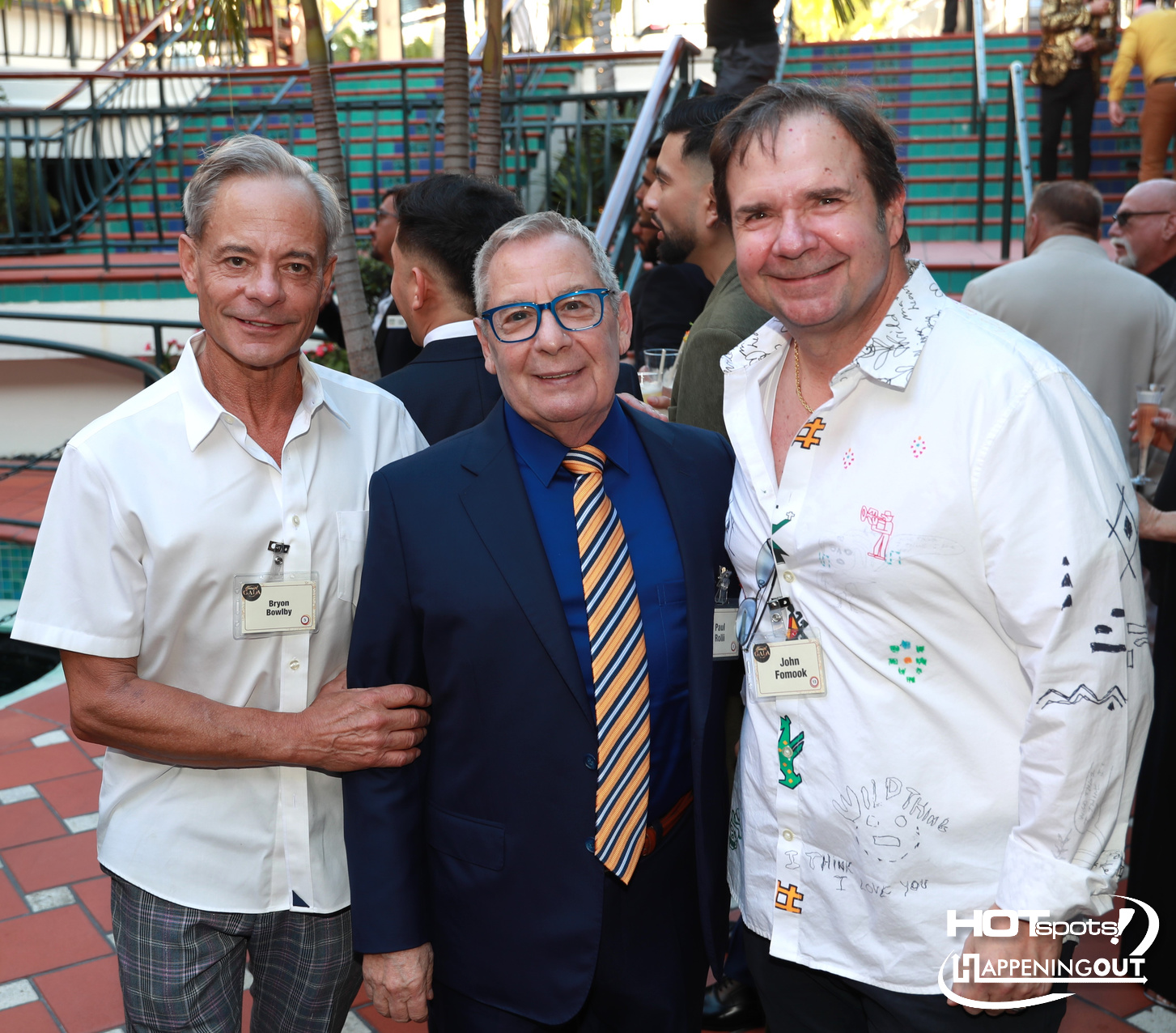 Three men pose for a photo at an outdoor event; left in a white shirt, middle in a navy suit with orange striped tie, right in a white patterned shirt, all wearing name badges.
