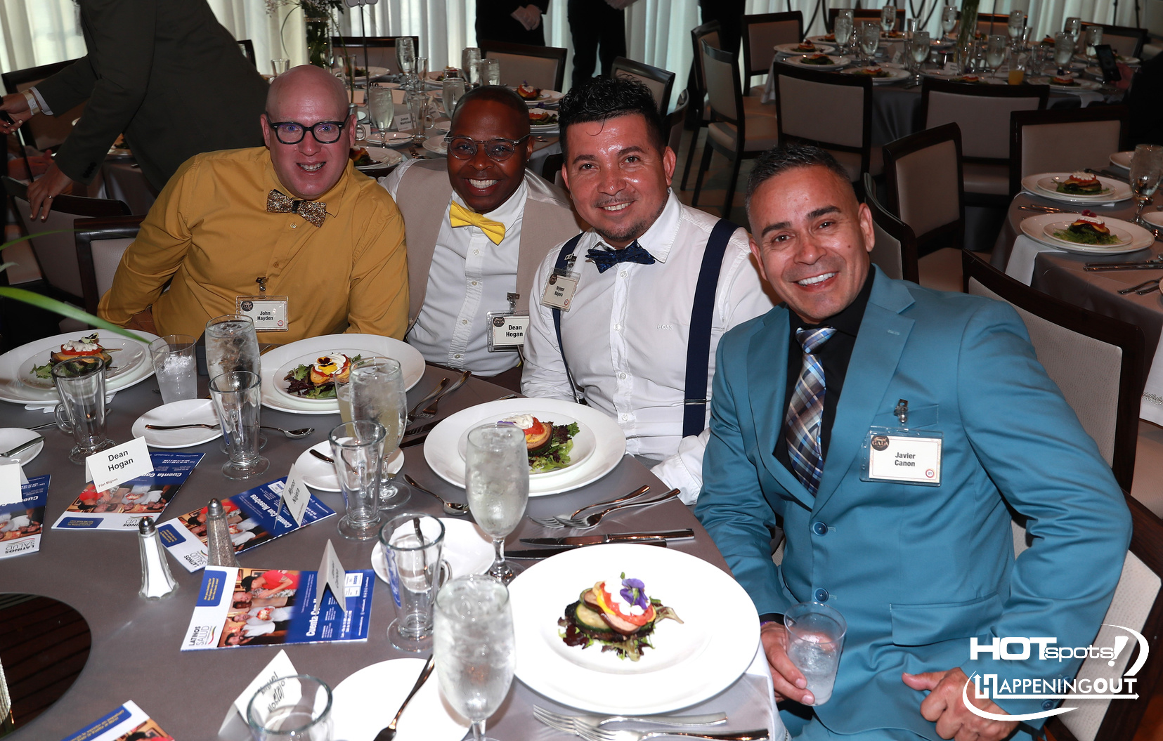 Four men in colorful formal attire sit at a round table during a banquet, smiling at the camera with plated salads in front of them.