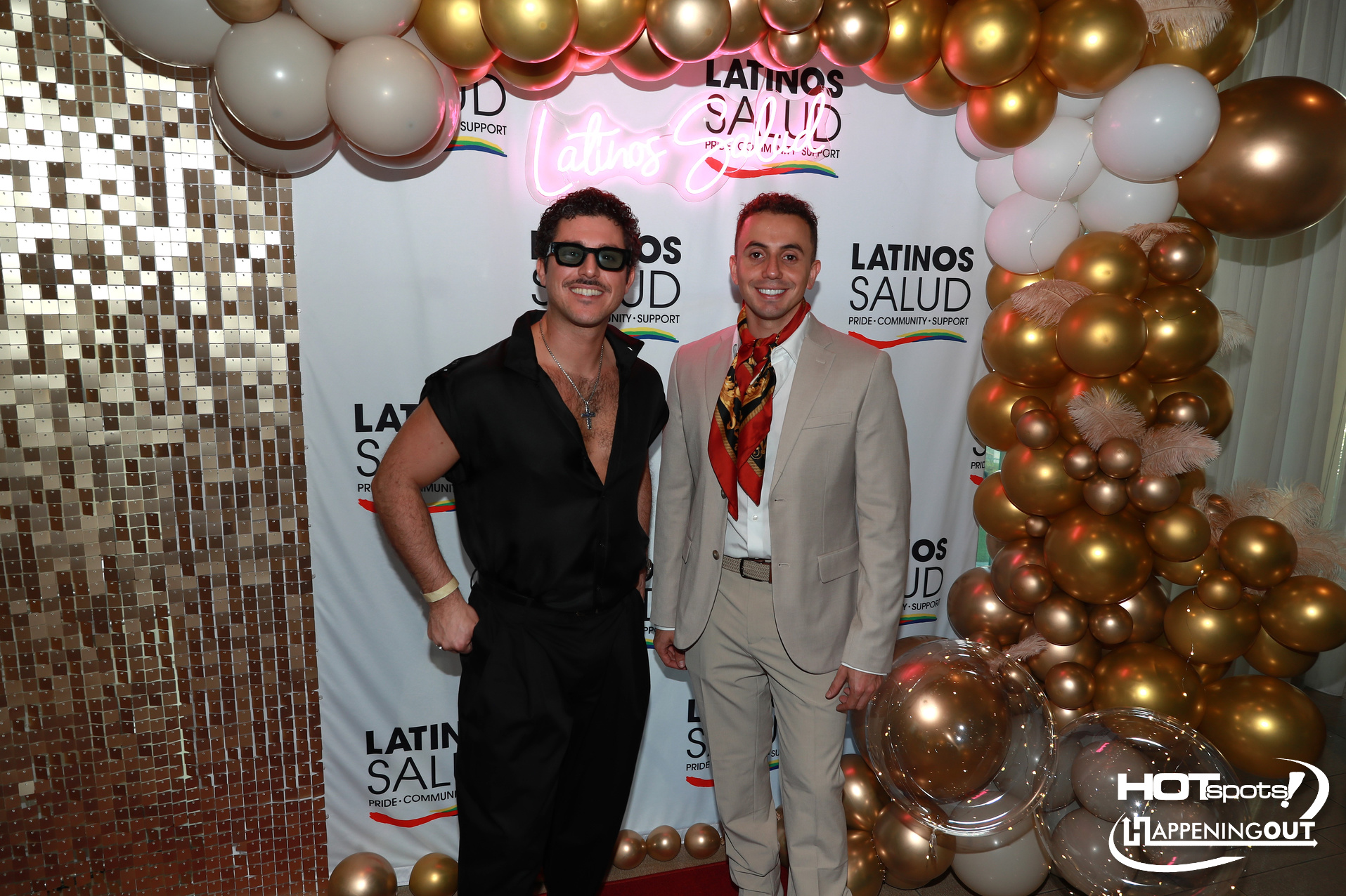 Two men pose and smile at a Latino Salud event, with a gold—and white balloon arch and banner backdrop reading 'LATINOS SALUD'.