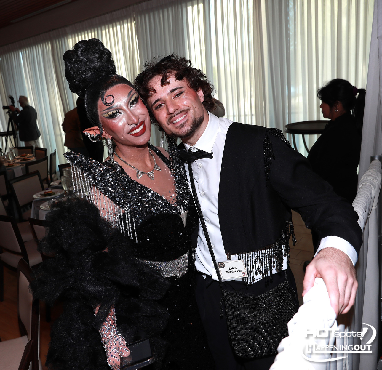 Two performers in glamorous outfits smile for the camera at an event, one in a sparkling black gown with dramatic makeup and the other in a white shirt with a black jacket and fringe details, posing together indoors.
