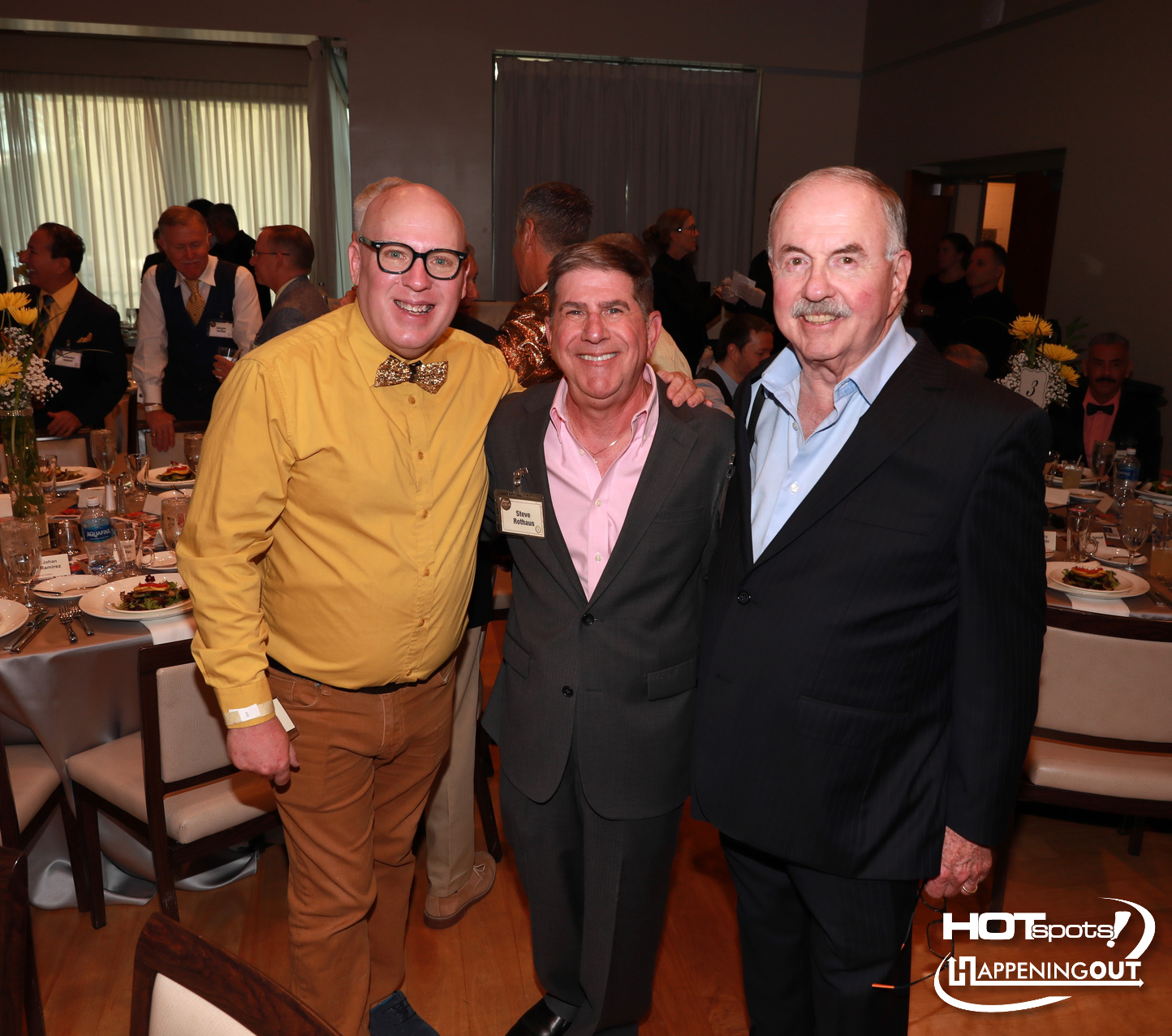 Three men in formal attire pose together at a banquet, smiling, with tables set for dinner in the background.