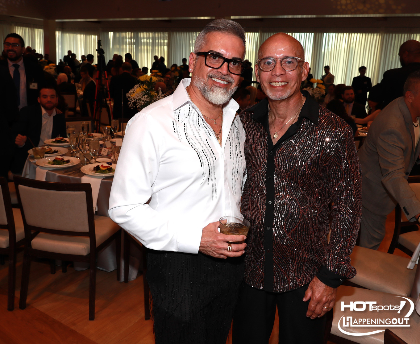 Two men in formal attire pose together at a banquet, one in a white embroidered shirt and the other in a sparkly jacket, holding drinks as guests dine in the background.