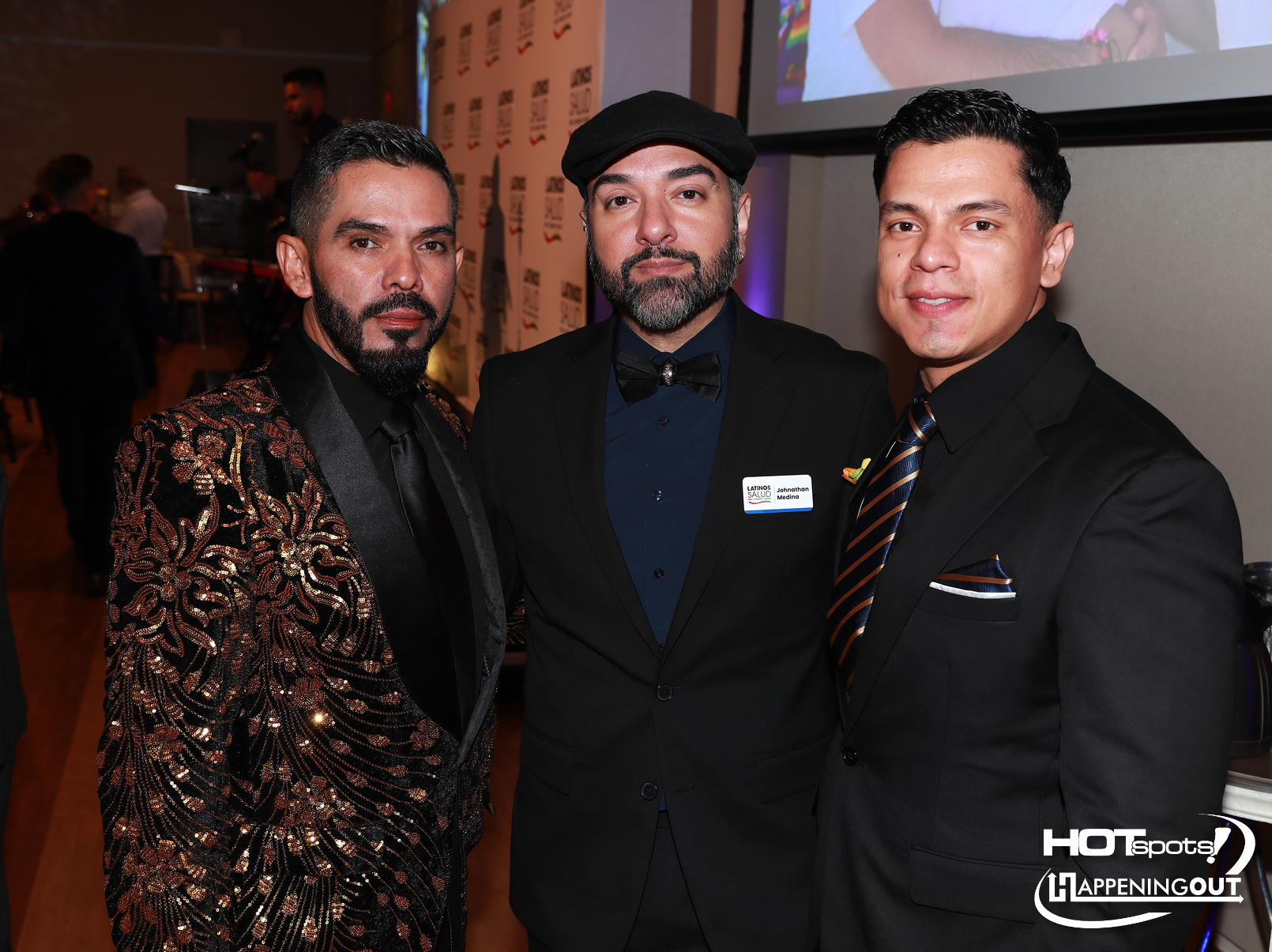 Three men in formal wear posing together at an event: left in a gold and black embroidered jacket, middle in a dark suit with bow tie and a cap, right in a black suit with a striped tie and pocket square, all smiling for the camera.
