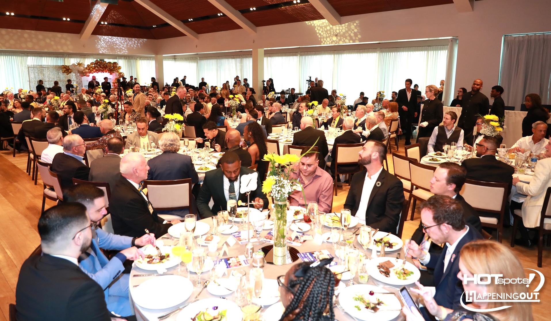 Banquet hall filled with round tables of formally dressed guests dining, yellow flower centerpieces and a balloon arch at the back stage area.