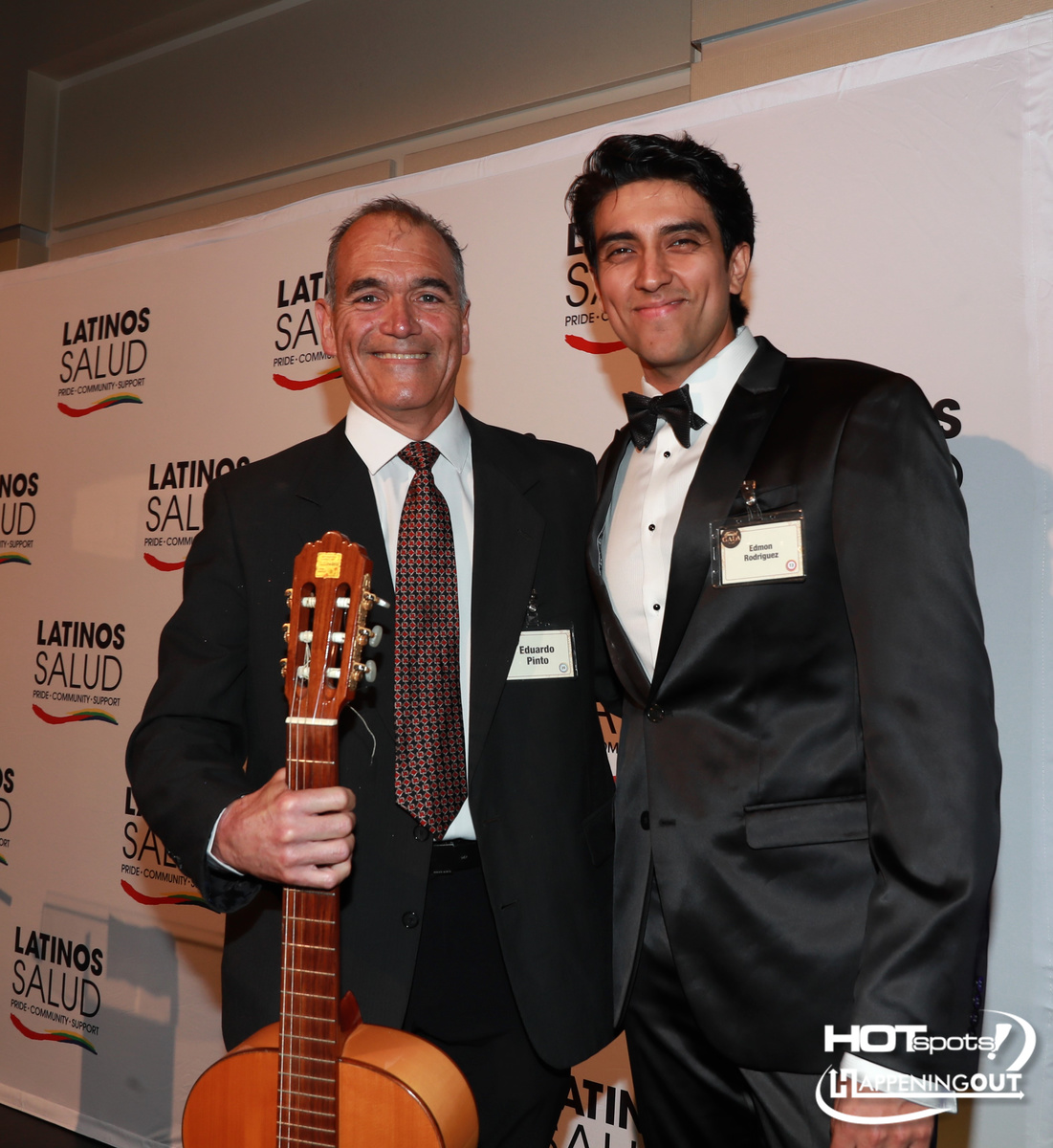 Two men in suits pose on a red carpet at a Latinos Salud event; one holds a guitar and smiles at the camera.