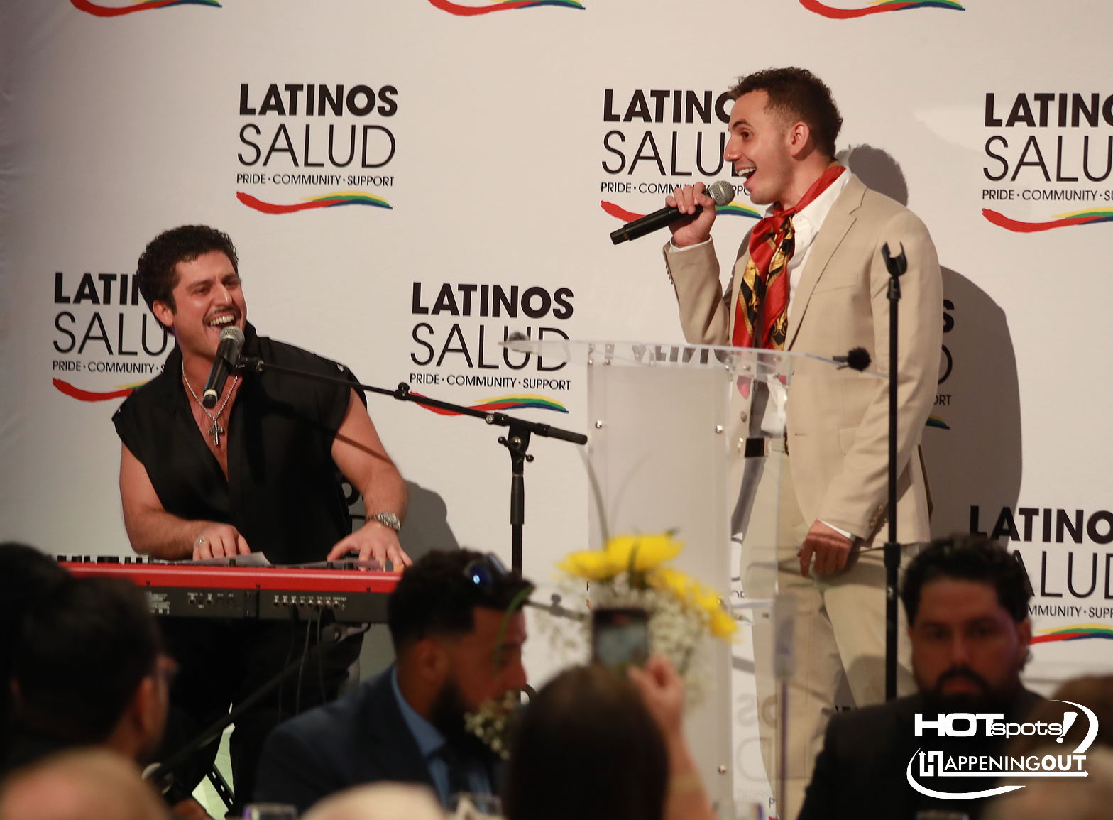 Two men perform on a Latinos Salud stage: one at a red keyboard singing, the other in a cream suit with a microphone at a podium.