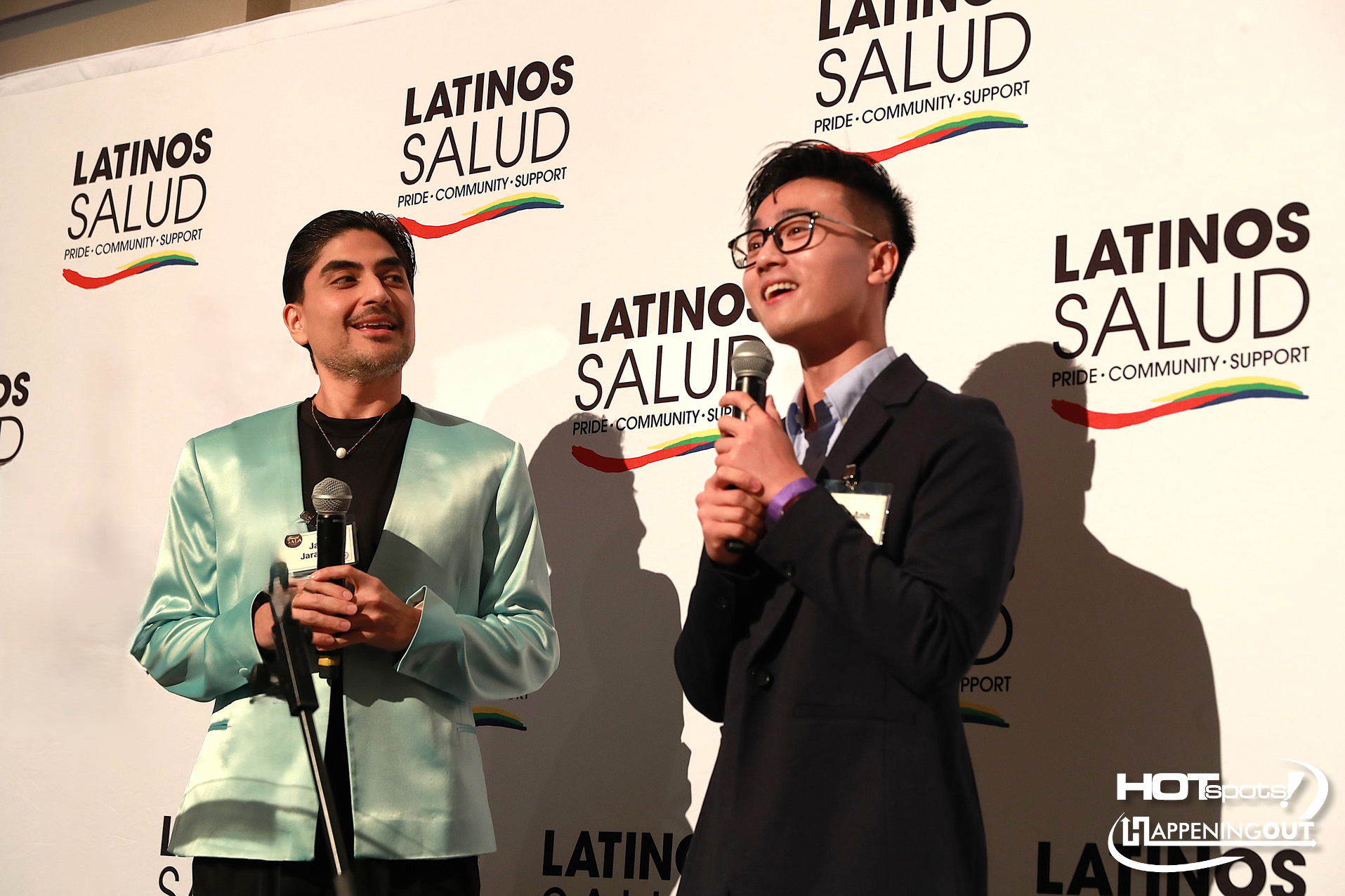 Two men in suits speak on a stage at a Latinos Salud event, each holding a microphone in front of a branded backdrop with the organization's logo and a rainbow accent.