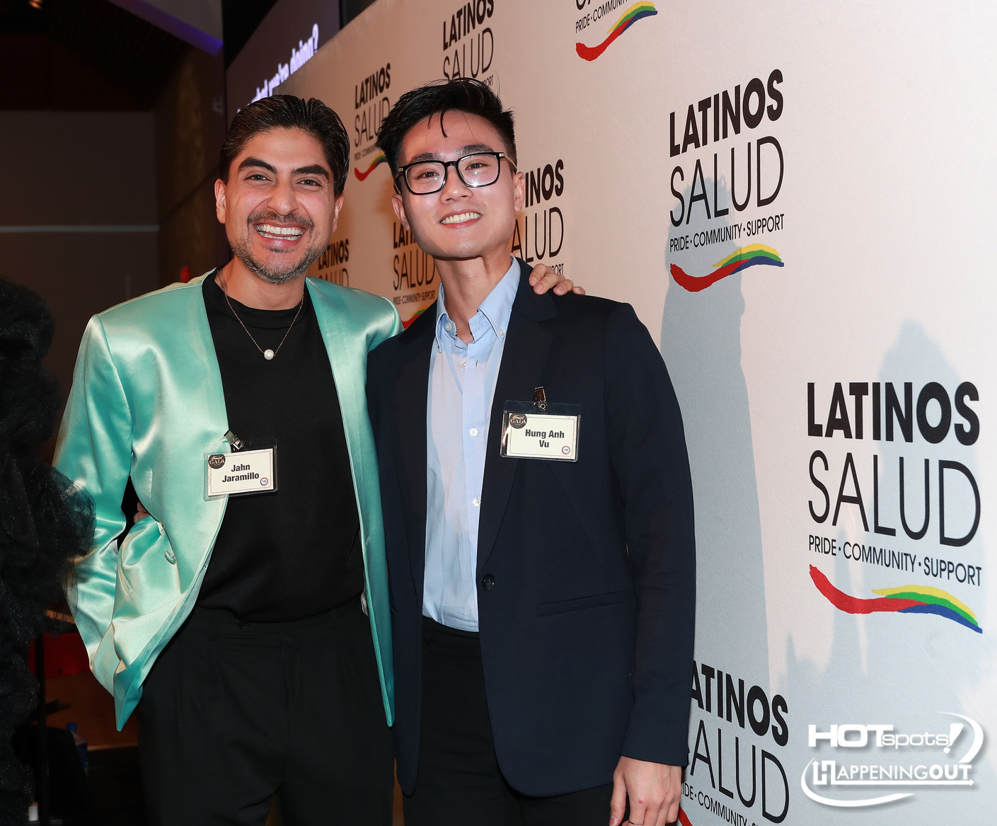 Two men pose together at a Latinos Salud event; one in a turquoise satin jacket and black shirt, the other in a navy blazer and glasses, both with name badges, against a Latinos Salud backdrop.