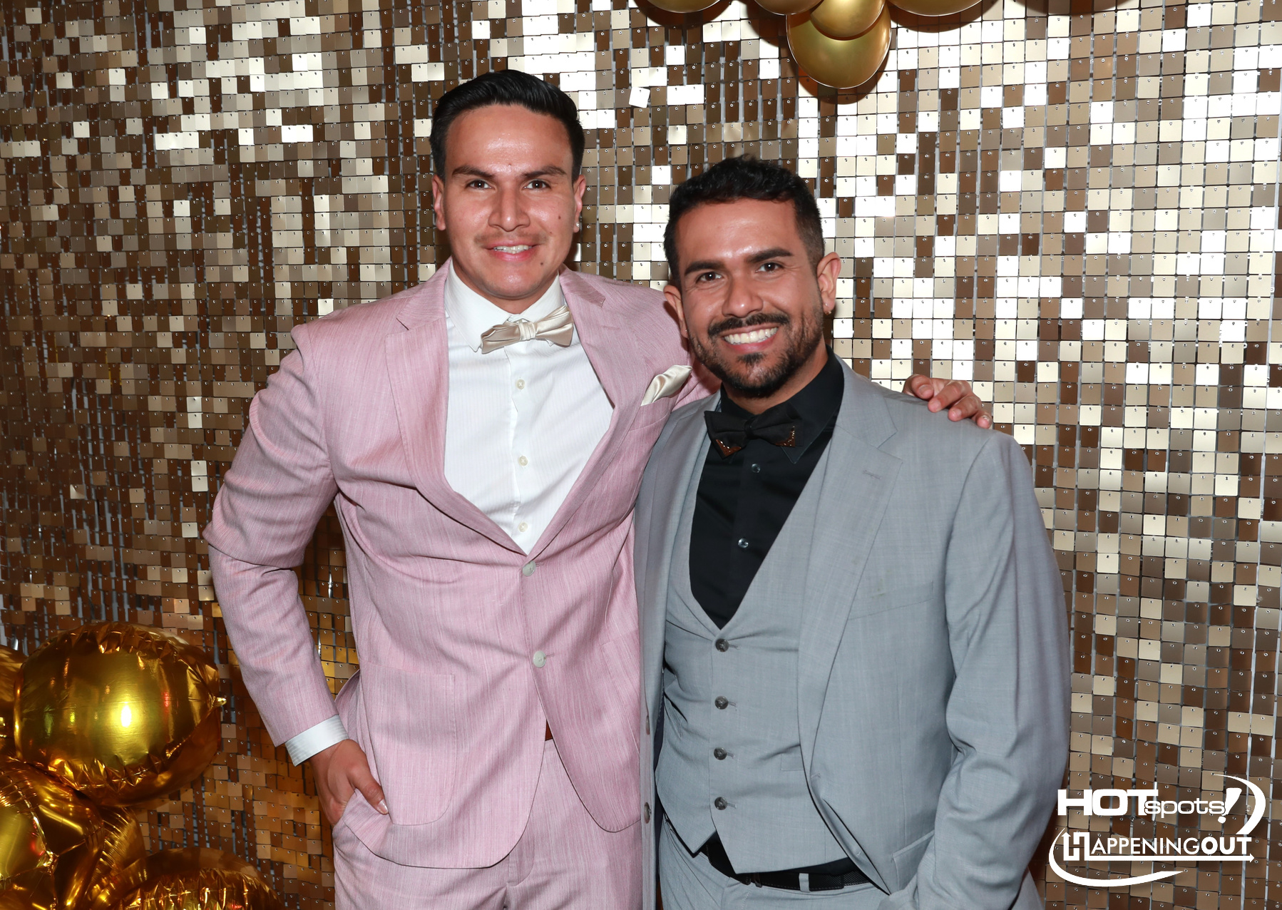 Two men in suits pose together at a festive photo booth with a gold sequin backdrop; one wears a pink suit with a cream bow tie, the other a gray suit with a black shirt and bow tie, both smiling.