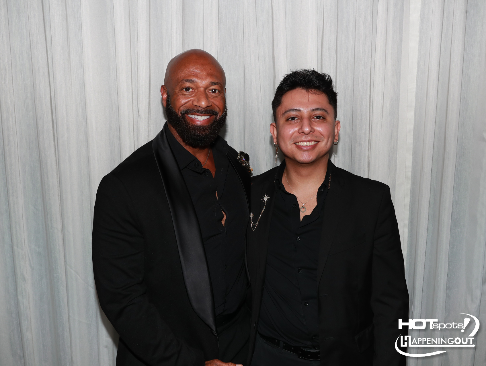 Two men in black suits posing together and smiling in front of a light gray backdrop; event logos appear bottom right corner.