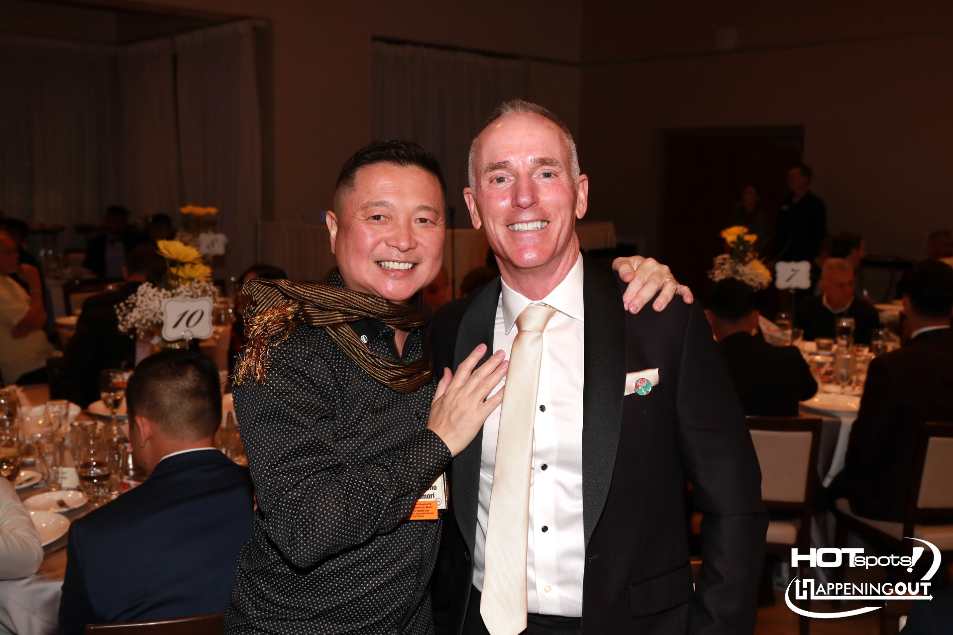 Two people smile for a photo at a formal banquet, with tables set for dinner in the background.