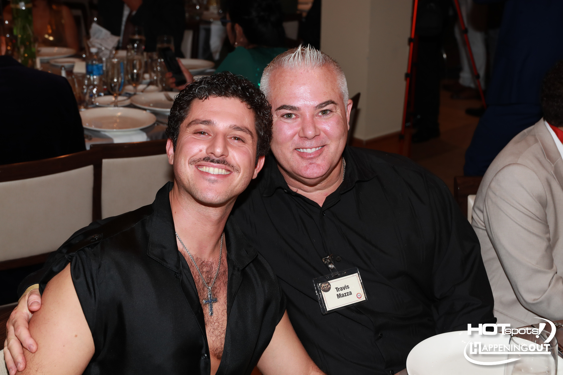 Two men in black shirts smiling at a banquet table during a formal dinner; one man wears a name badge reading Travis Mazza in front.