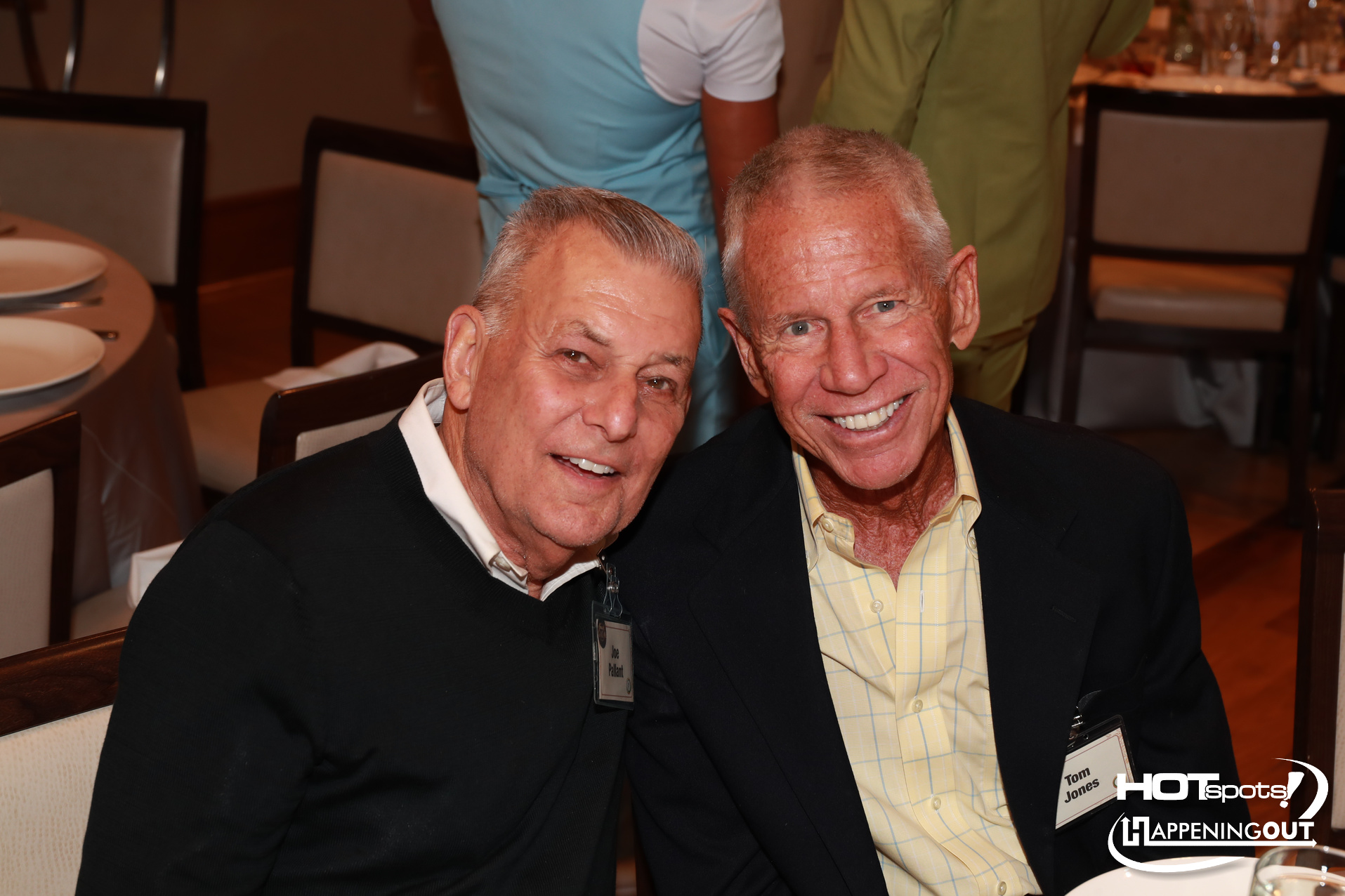 Two senior men in dark jackets lean together and smile at the camera during a formal dinner event at a banquet table with place settings nearby.