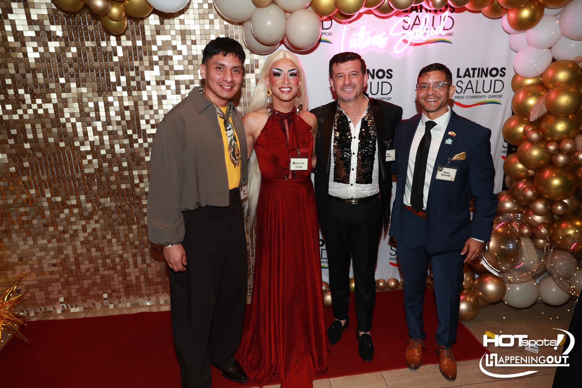 Four people pose on a red carpet with balloons and a gold mosaic backdrop; a drag queen in a red gown stands with three men in suits, smiling.