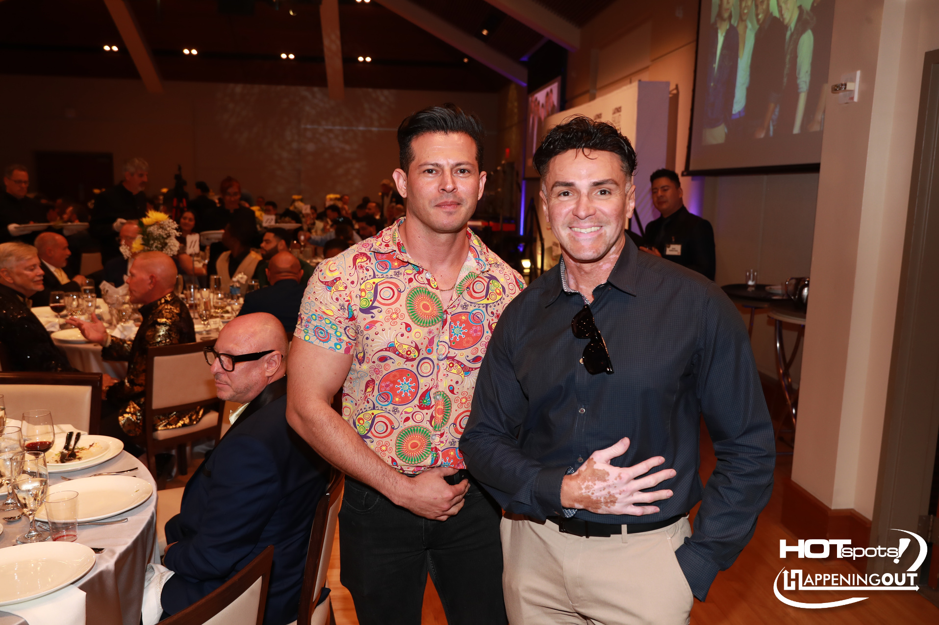 Two men posing for a photo at a formal banquet, one in a colorful paisley shirt and the other in a dark blue shirt, with guests and a projector screen in the background.