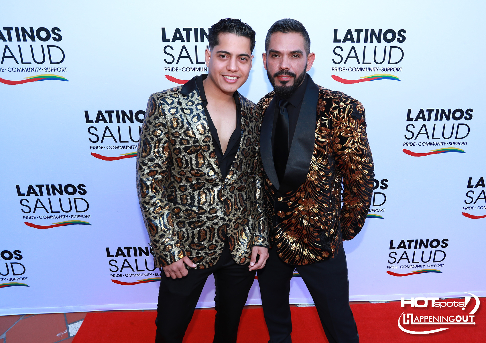 Two men in sequined jackets pose together on a red carpet at a Latinos Salud event, smiling for the camera with a branded backdrop behind them.