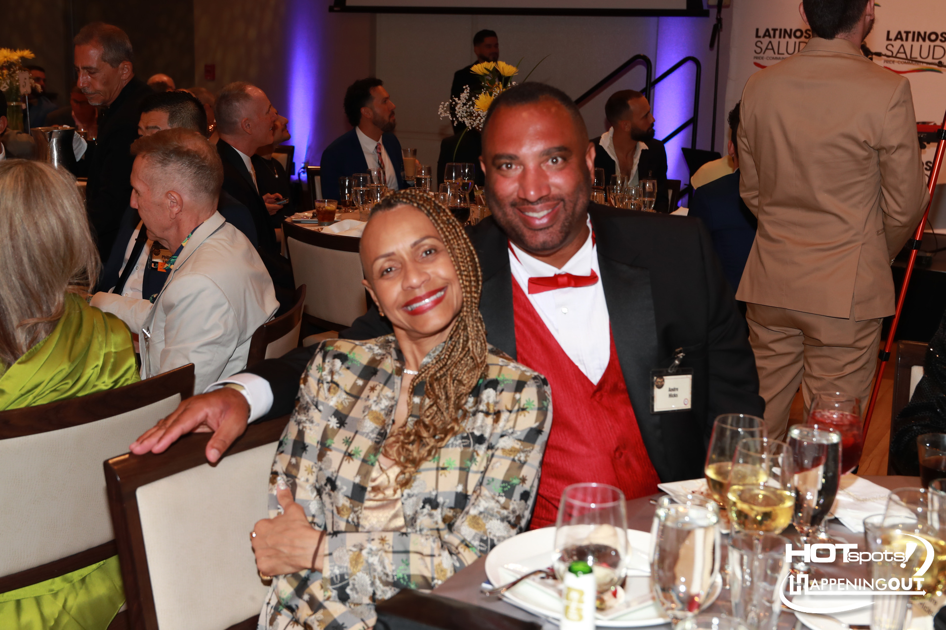 Smiling couple at a banquet table during a formal event, woman in a patterned blazer and man in a red bow tie with name badge visible on his lapel.