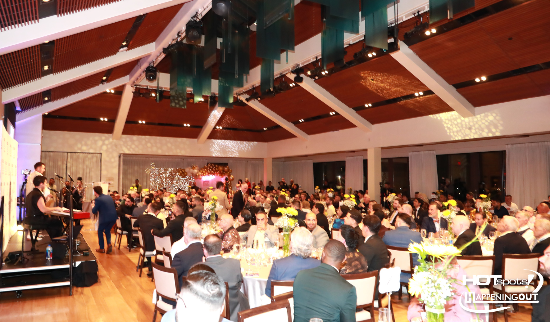 A large formal dinner in a modern hall with a live band on stage and many guests seated at round tables with centerpieces and yellow flowers, soft stage lighting visible.