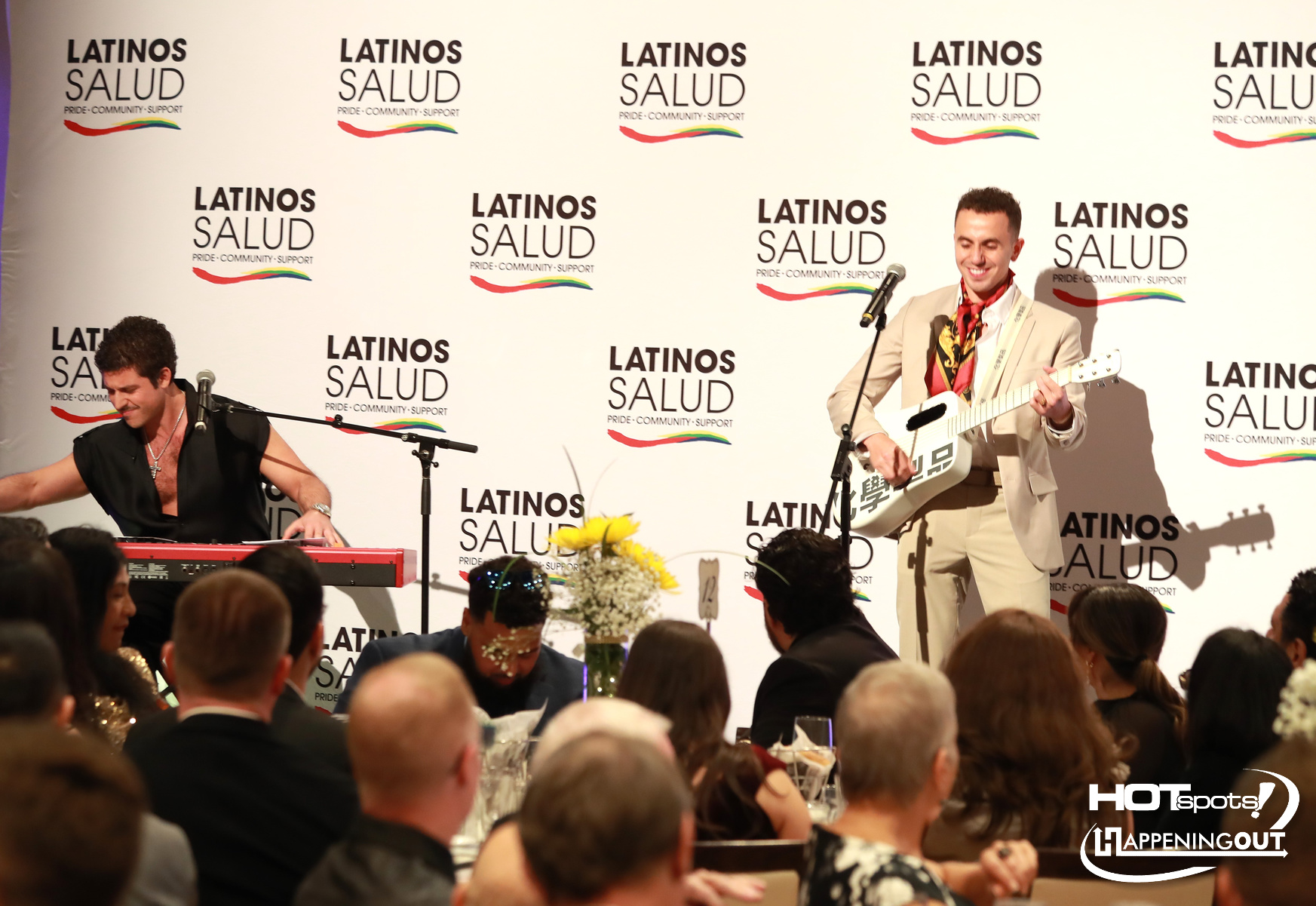 Two musicians perform on a stage at a Latinos Salud event; a keyboard player on the left and a guitarist on the right, with an audience seated in front and a Latinos Salud backdrop behind them.