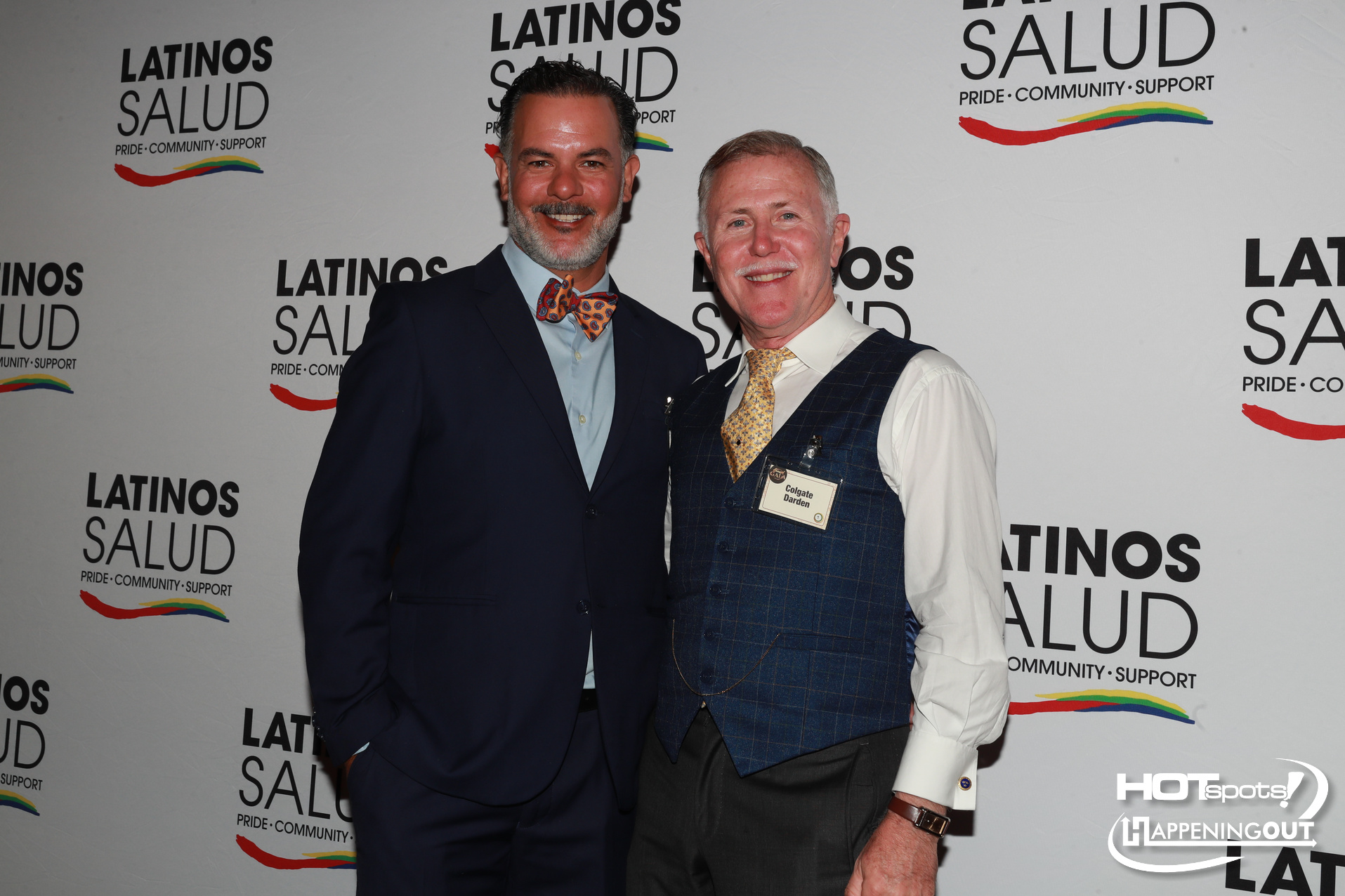Two men posing at a Latino Salud event; one in a navy suit with an orange bow tie, the other in a blue checkered vest and yellow tie, name tag reads Colgate Darden, backdrop reads LATINOS SALUD with a rainbow stripe.