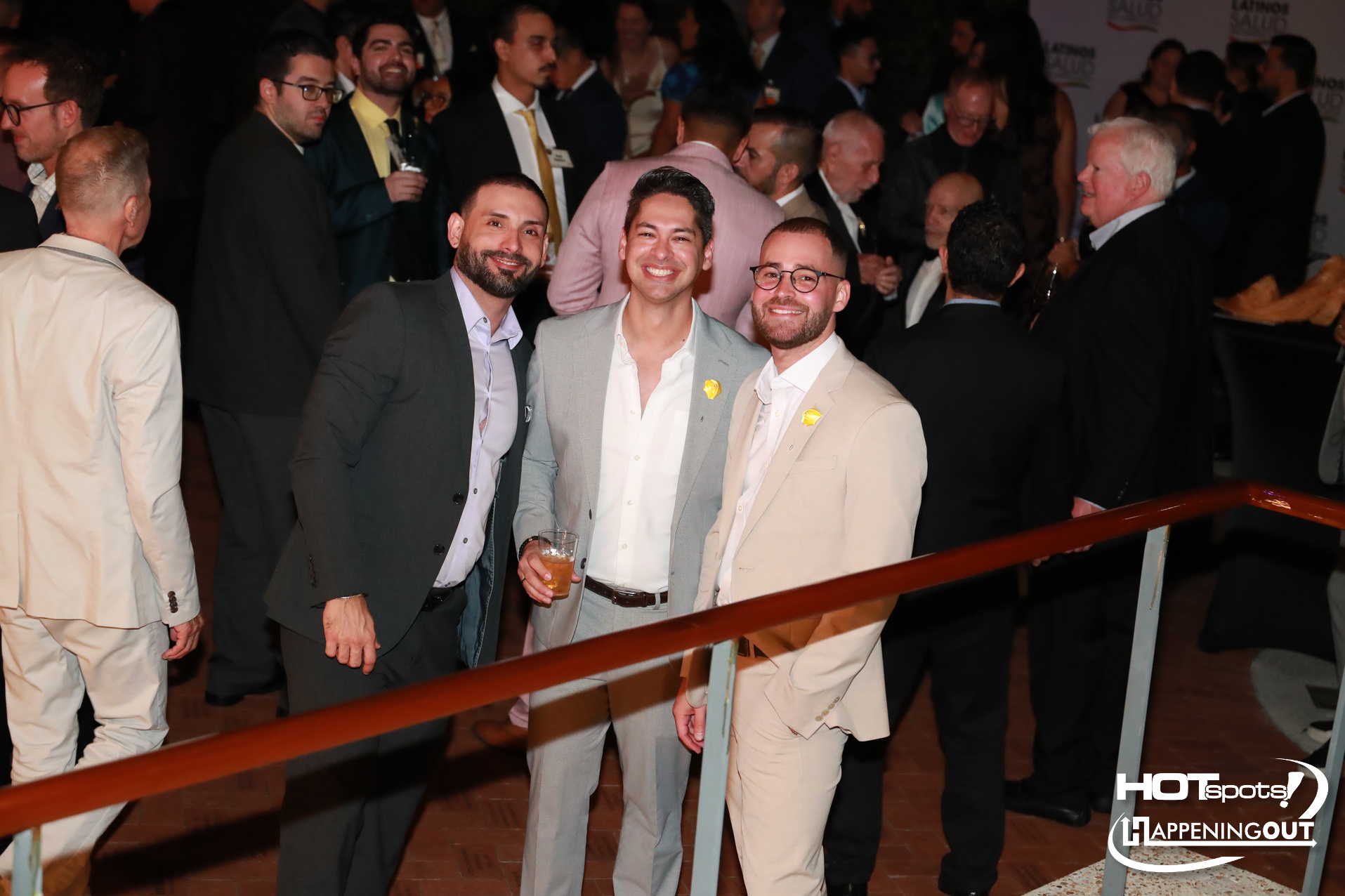 Three men in light suits smile at the camera at a crowded formal event; one holds a drink, with a logo visible in the bottom-right corner.