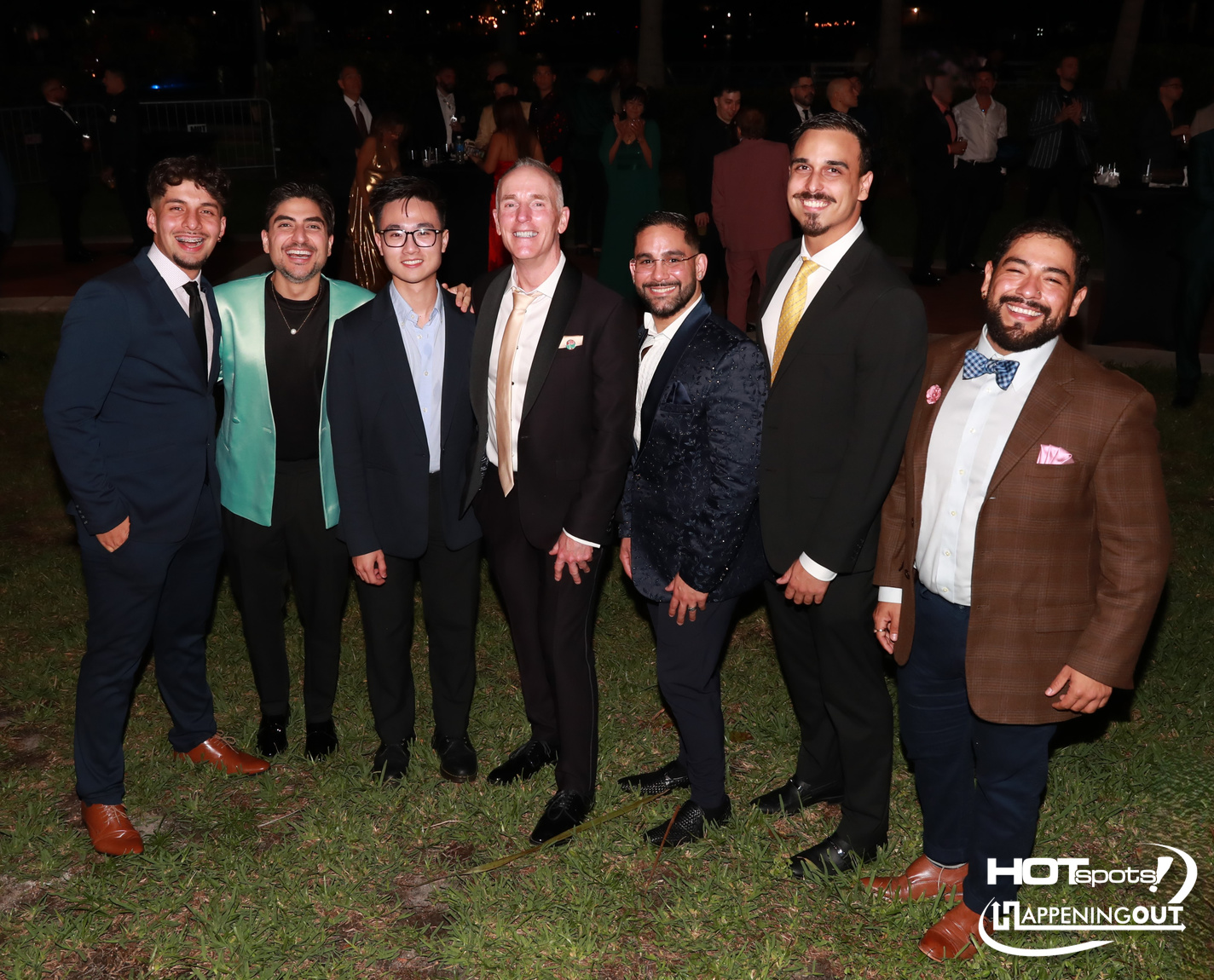 Seven men in suits posing together at an outdoor night event, smiling on a grassy area.