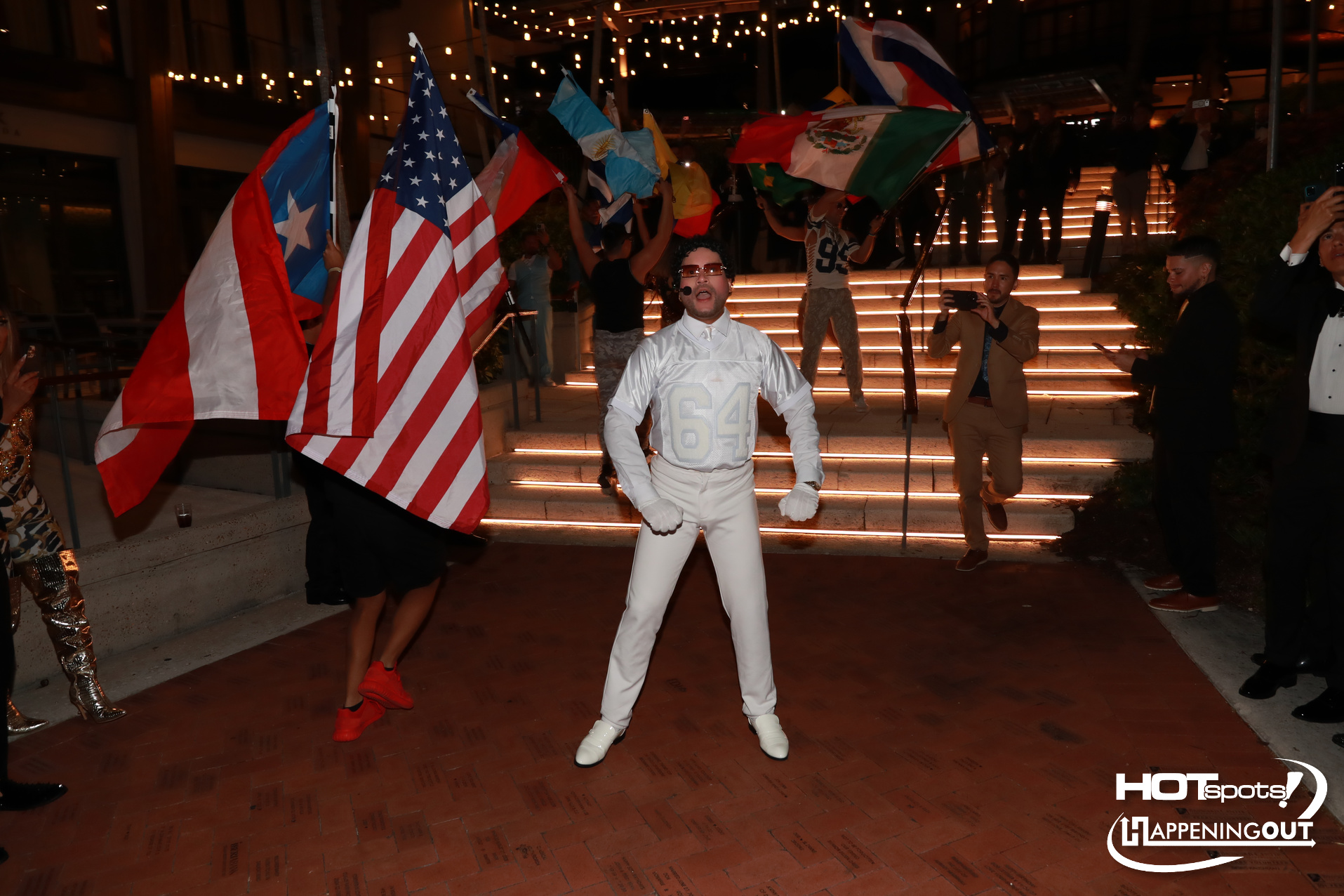 Dancer in a white numbered suit (64) performing on lit steps while flag-waving crowd watches at a night event with string lights.