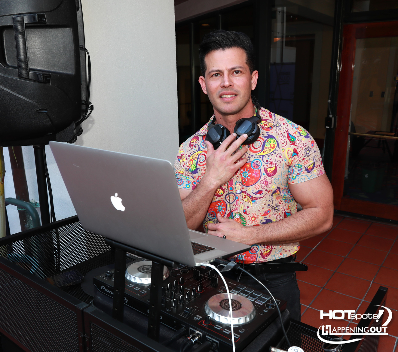 DJ in a colorful patterned shirt stands behind a laptop and mixer with headphones around his neck at a party setup